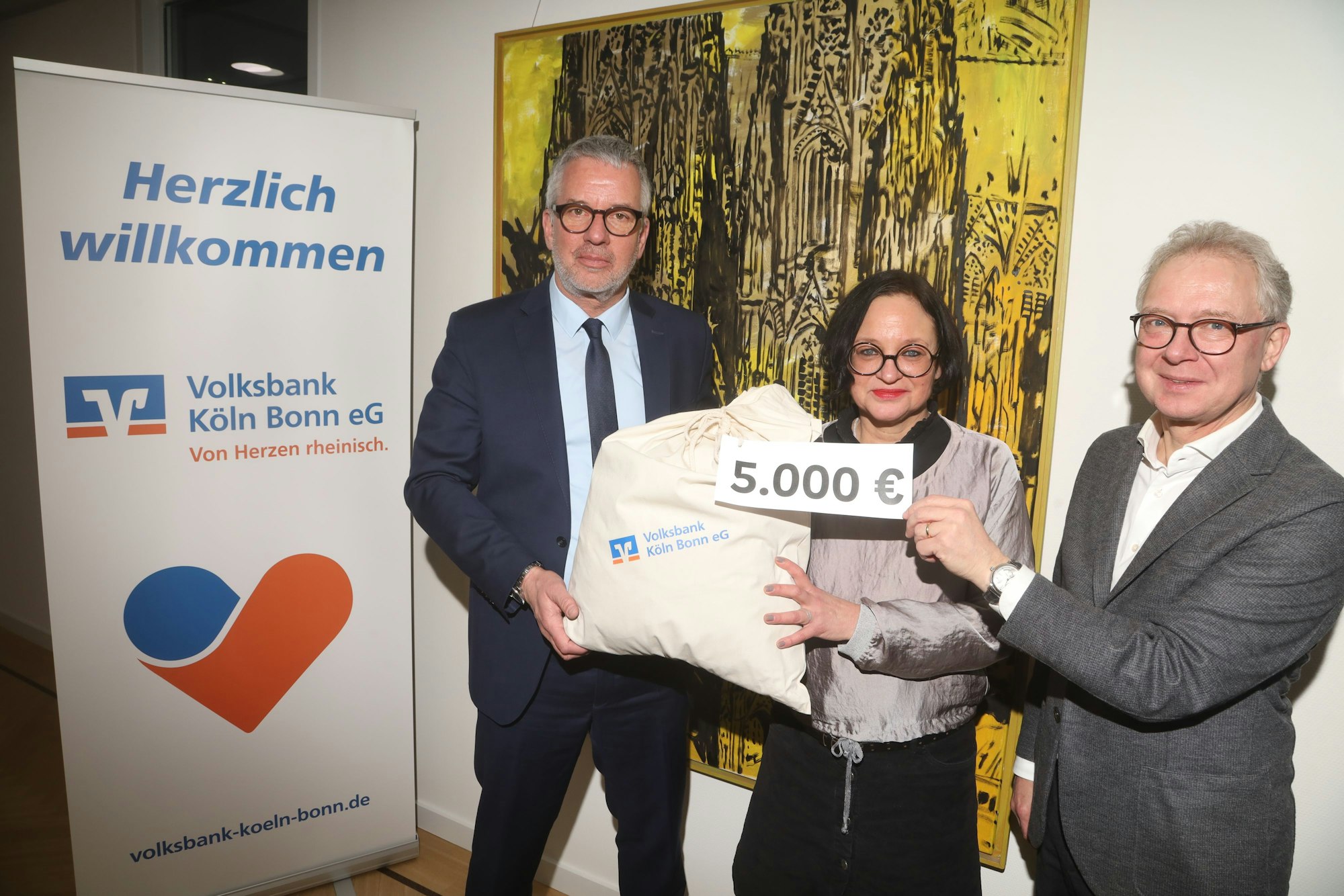 Jürgen Pütz, Vorstandsvorsitzender der Volksbank Köln Bonn, überreicht vor einem modernen Gemälde des Kölner Doms Christian Hümmeler und Caroline Kron vom „wir helfen“-Vorstandsteam einen symbolischen Spendensack über 5.000 Euro.