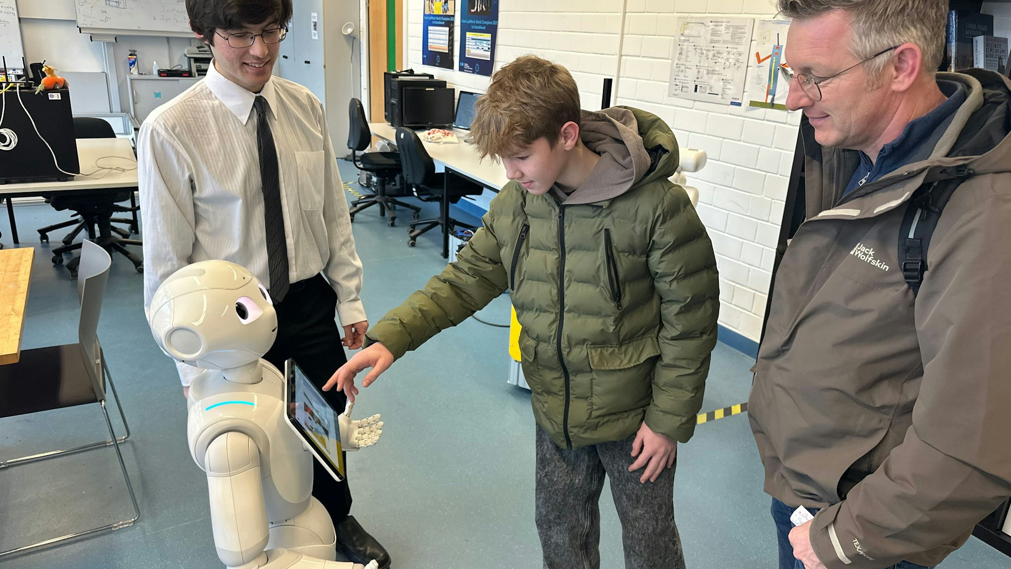 Der 14-jährige Laron Fuchs (M.) ist mit seinem Vater Andreas (r.) aus Windeck gekommen, um sich über Studienmöglichkeiten zu informieren. Jordan Schneider (l.), wissenschaftlicher Mitarbeiter der Hochschule, erklärt ihm die Funktionsweise eines sozialen Roboters.