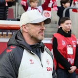 Lukas Kwasniok trägt eine weiße Jacke und eine weiße Baseballmütze des 1. FC Köln und blickt aufs Spielfeld.