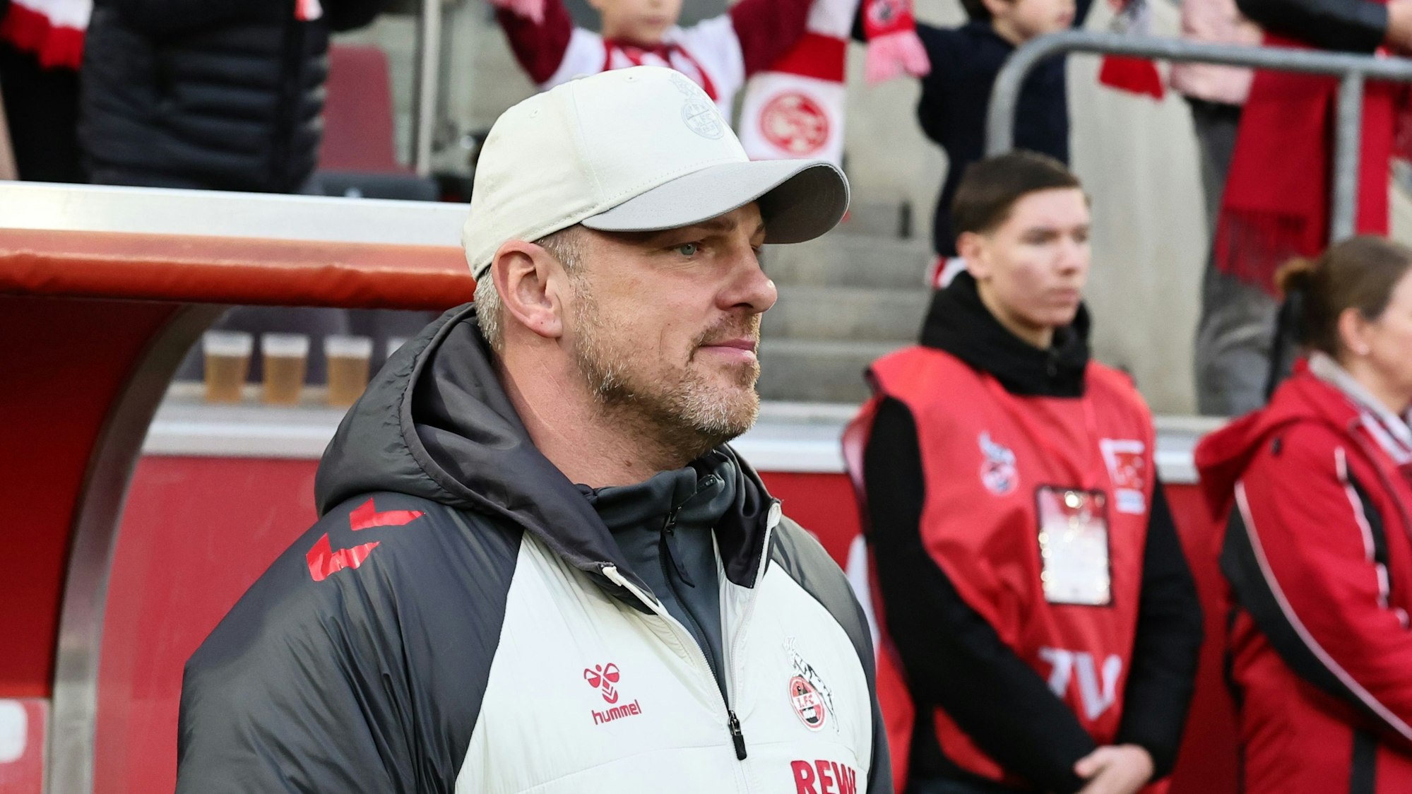 Lukas Kwasniok trägt eine weiße Jacke und eine weiße Baseballmütze des 1. FC Köln und blickt aufs Spielfeld.