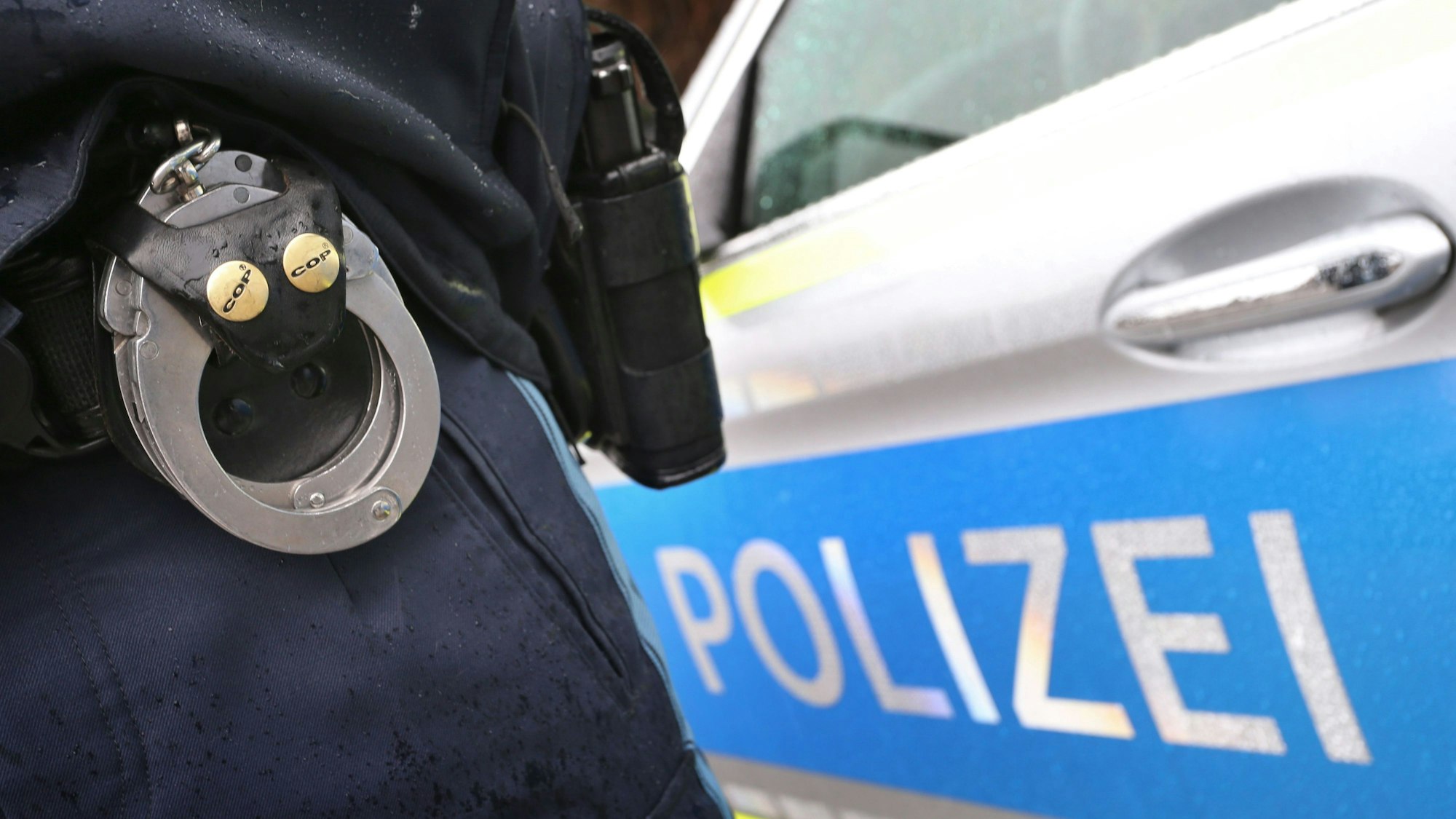 Ein Polizist mit Handschellen und einer Pistole am Gürtel steht vor einem Streifenwagen.