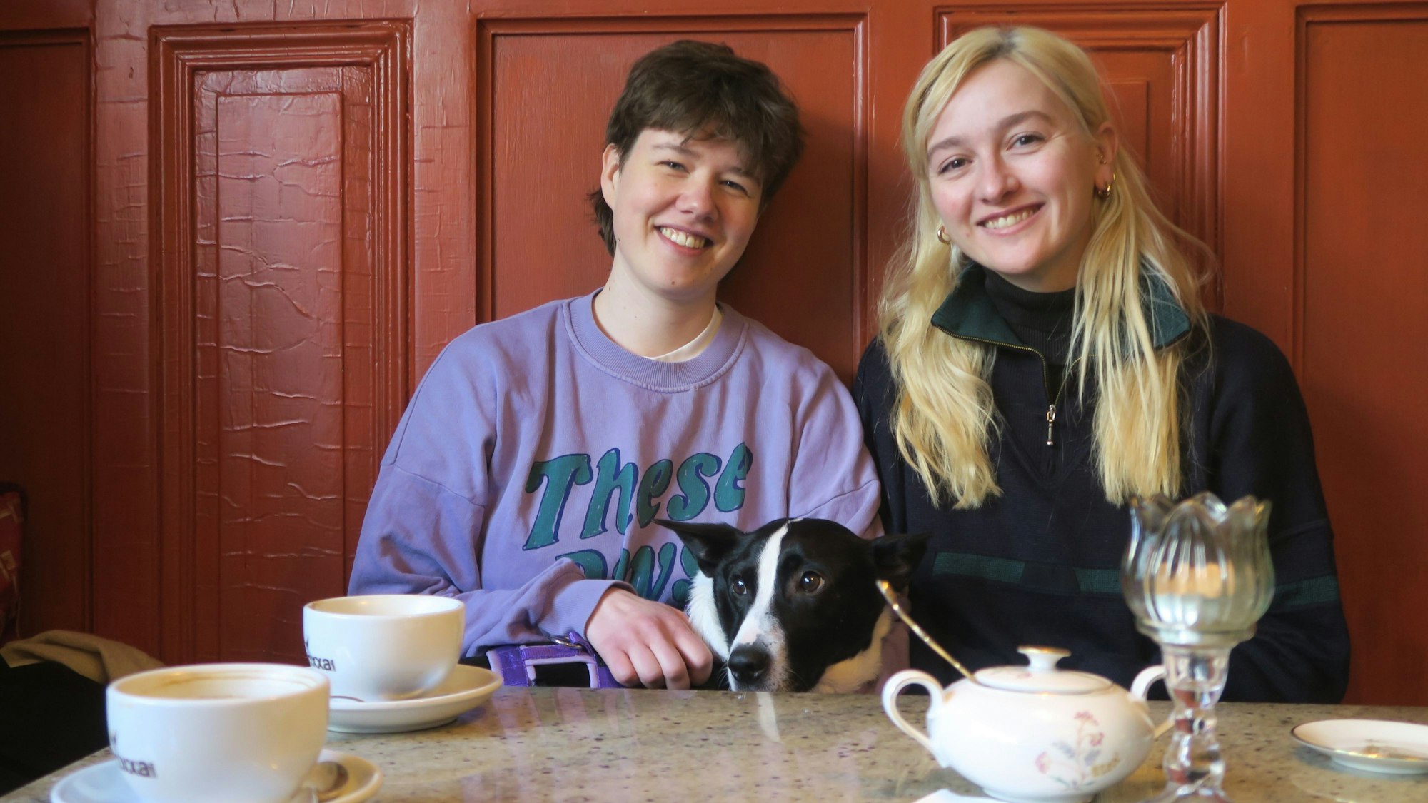 Zwei Frauen sitzen an einem Café-Tisch, zwischen ihnen lugt der schwarz-weiße Kopf eines Hundes über die Tischplatte.