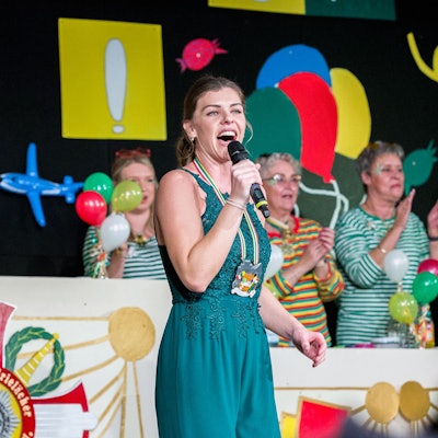 Eine Frau im blauen Kleid steht singend mit Mikrofon in der Hand auf einer Bühne.