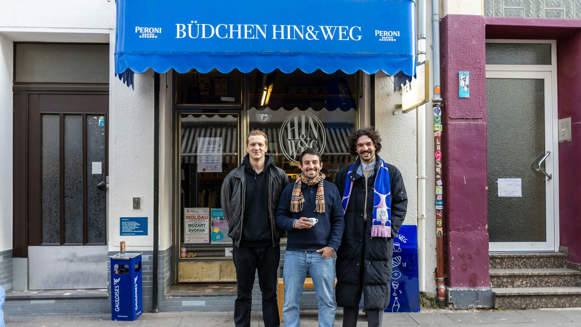 Drei Männer stehen vor einem Laden und lächeln in die Kamera.