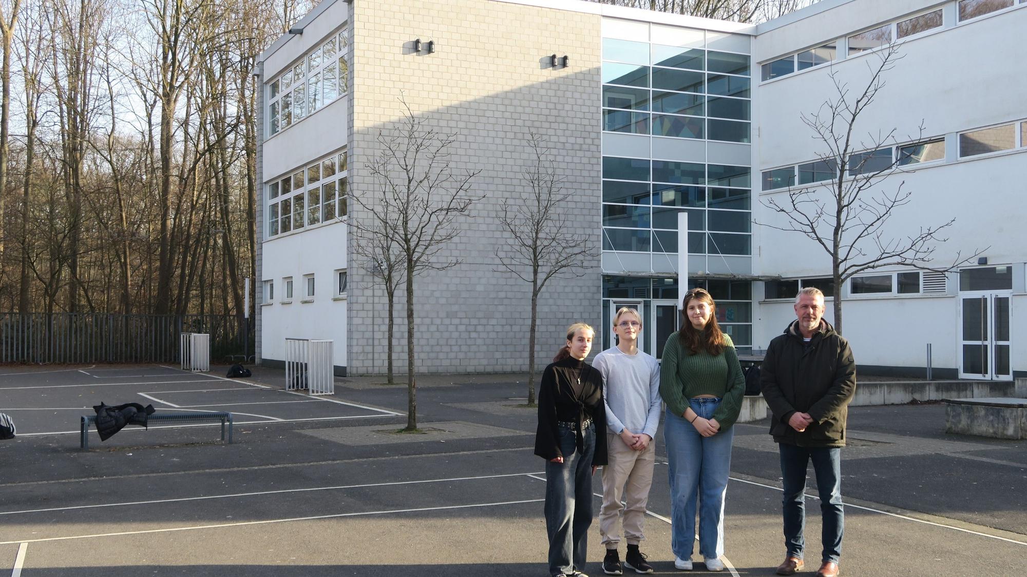 Drei Schüler und ein Lehrer stehen auf einem großen asphaltierten Schulhof.