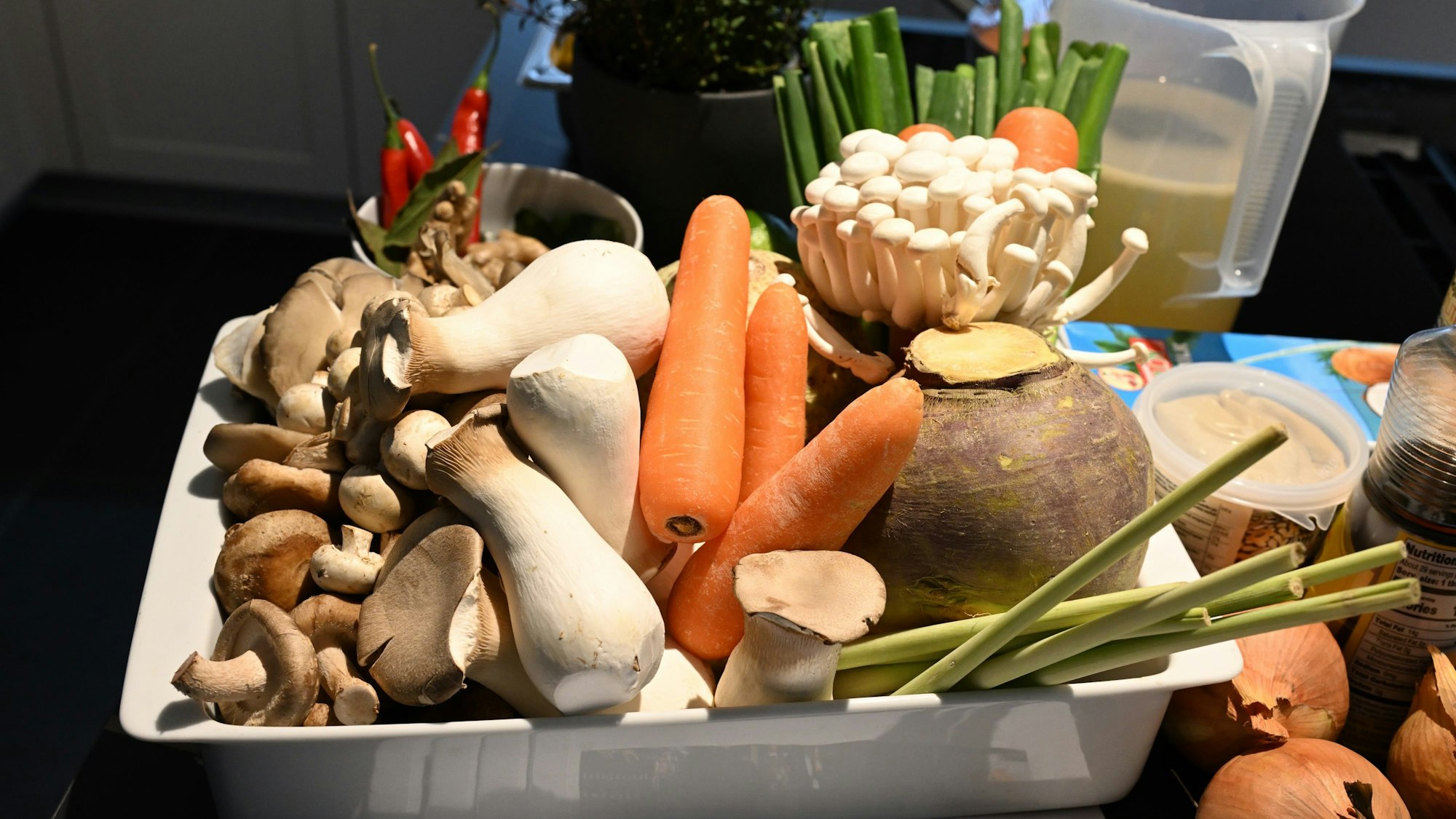 Verschiedenes Gemüse, darunter Möhren, Pilze, Peperoni, Zwiebeln liegen in und neben einer Schale in einer Küche.