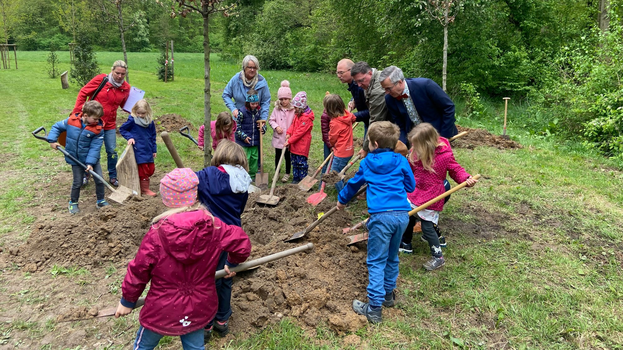 Viele Kinder und einige Erwachsene pflanzen mit Schaufeln bewaffnet einen jungen Baum ein.