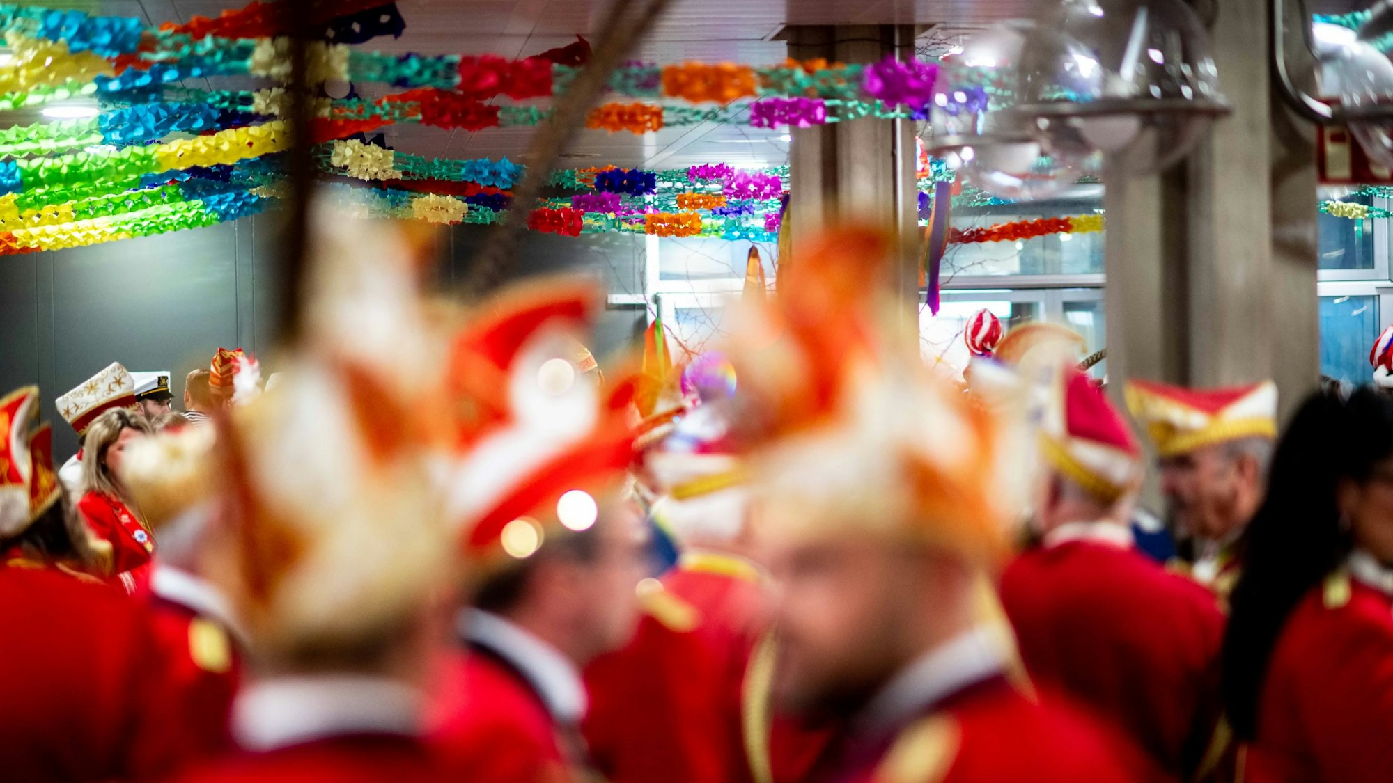 In einem mit Girlanden dekorierten Saal sind Karnevalisten mit roten Jacketts und Narrenkappen unscharf zu sehen.