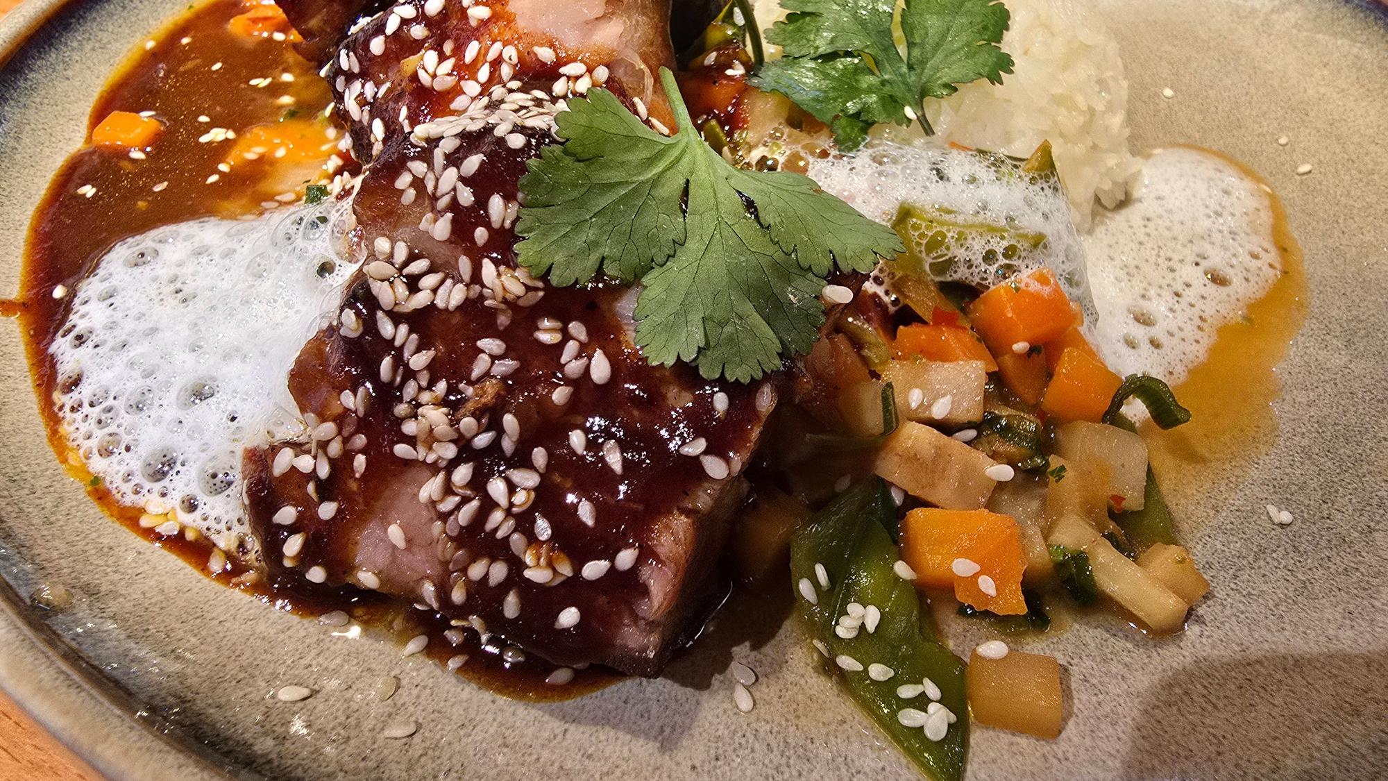 Auf einem hellen Teller liegt das Fleisch auf Gemüsebett und unter dunkelbrauner Sauce, bestreut mit Sesam.