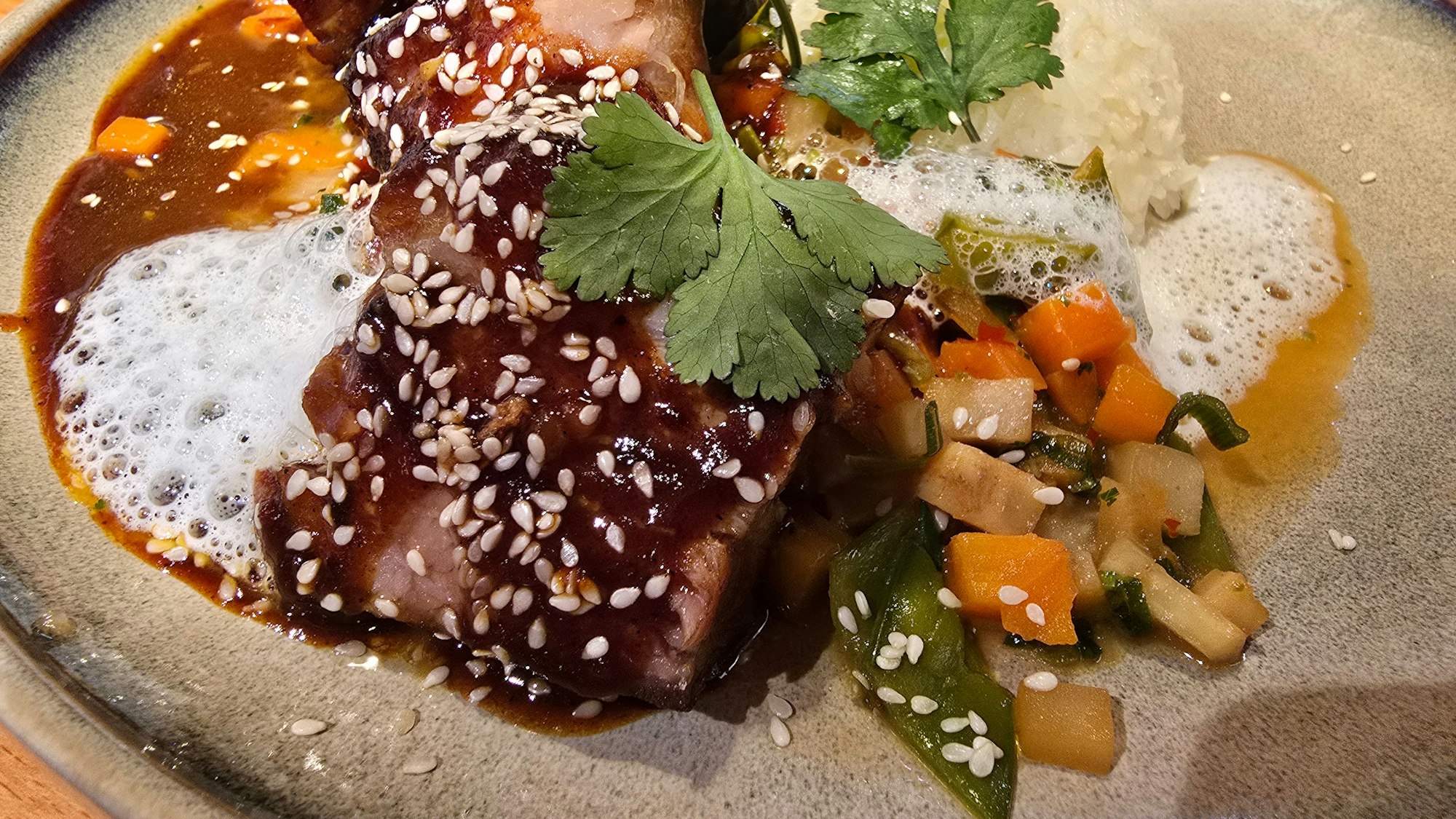 Auf einem hellen Teller liegt das Fleisch auf Gemüsebett und unter dunkelbrauner Sauce, bestreut mit Sesam.