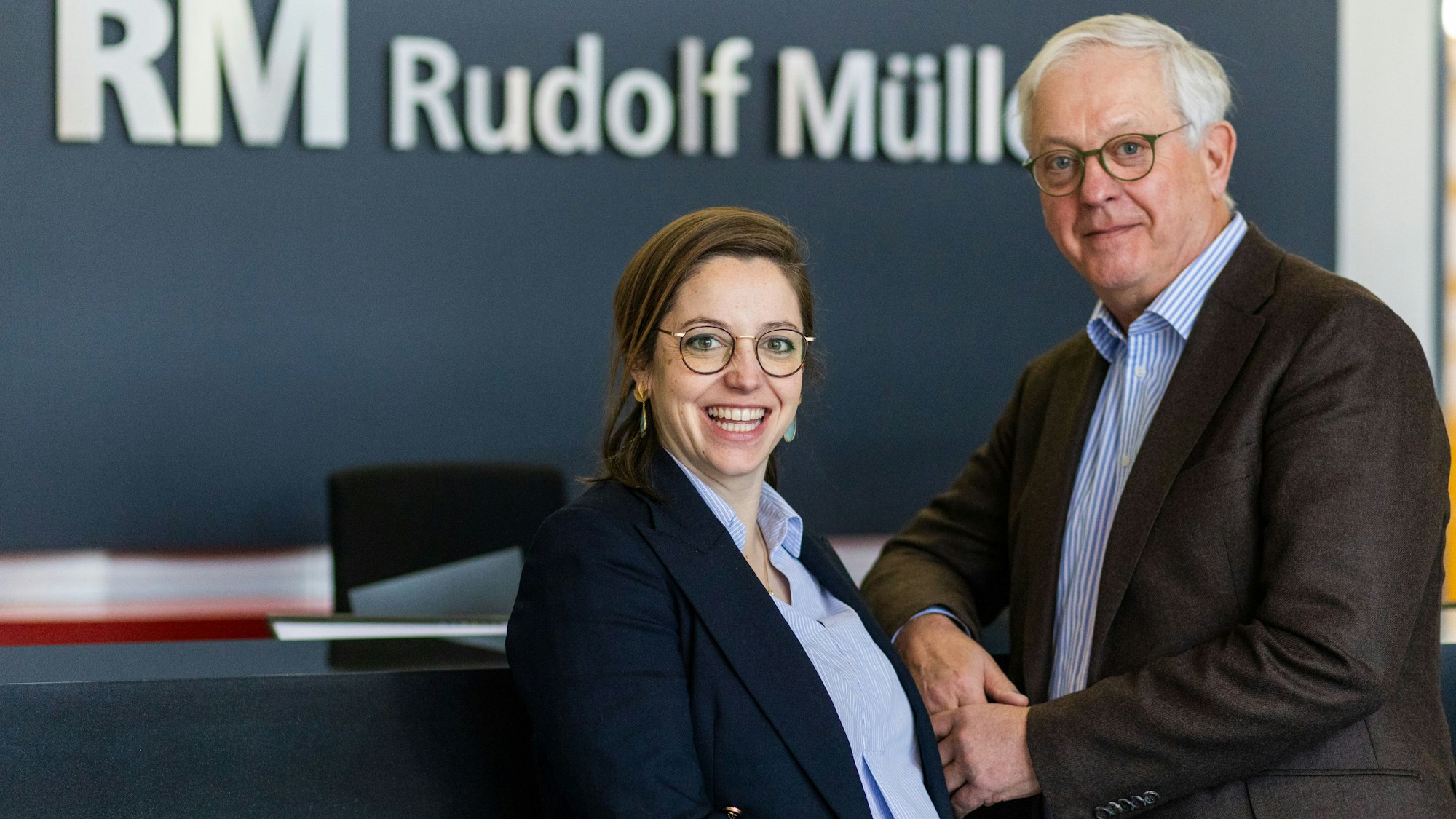 Eine jüngere Frau und ein älterer Mann lächeln in die Kamera.