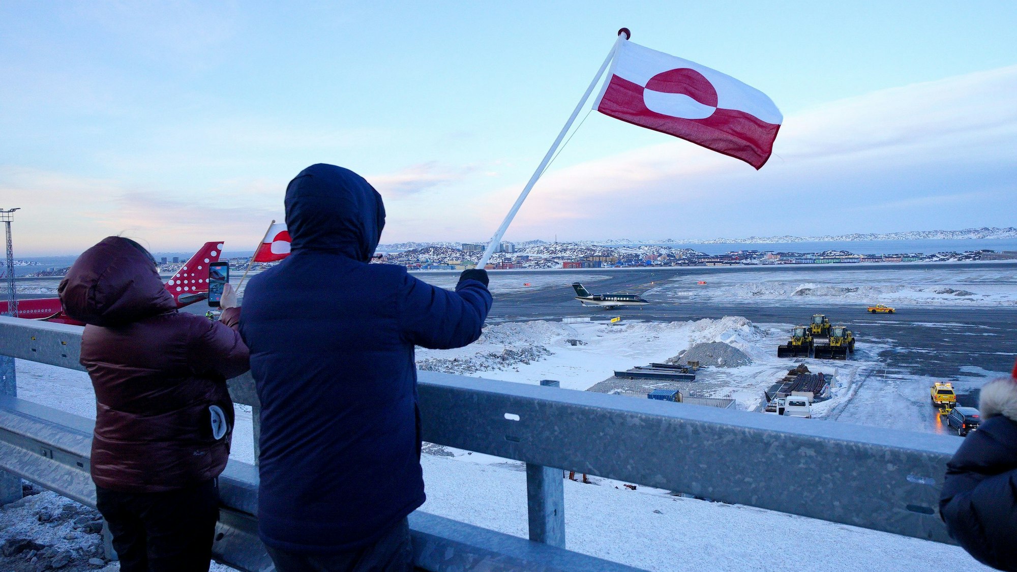 Das Foto zeigt Menschen mit der Flagge Grönlands am Flughafen von Nuuk. Foto: Ben Birchall/PA Wire/dpa