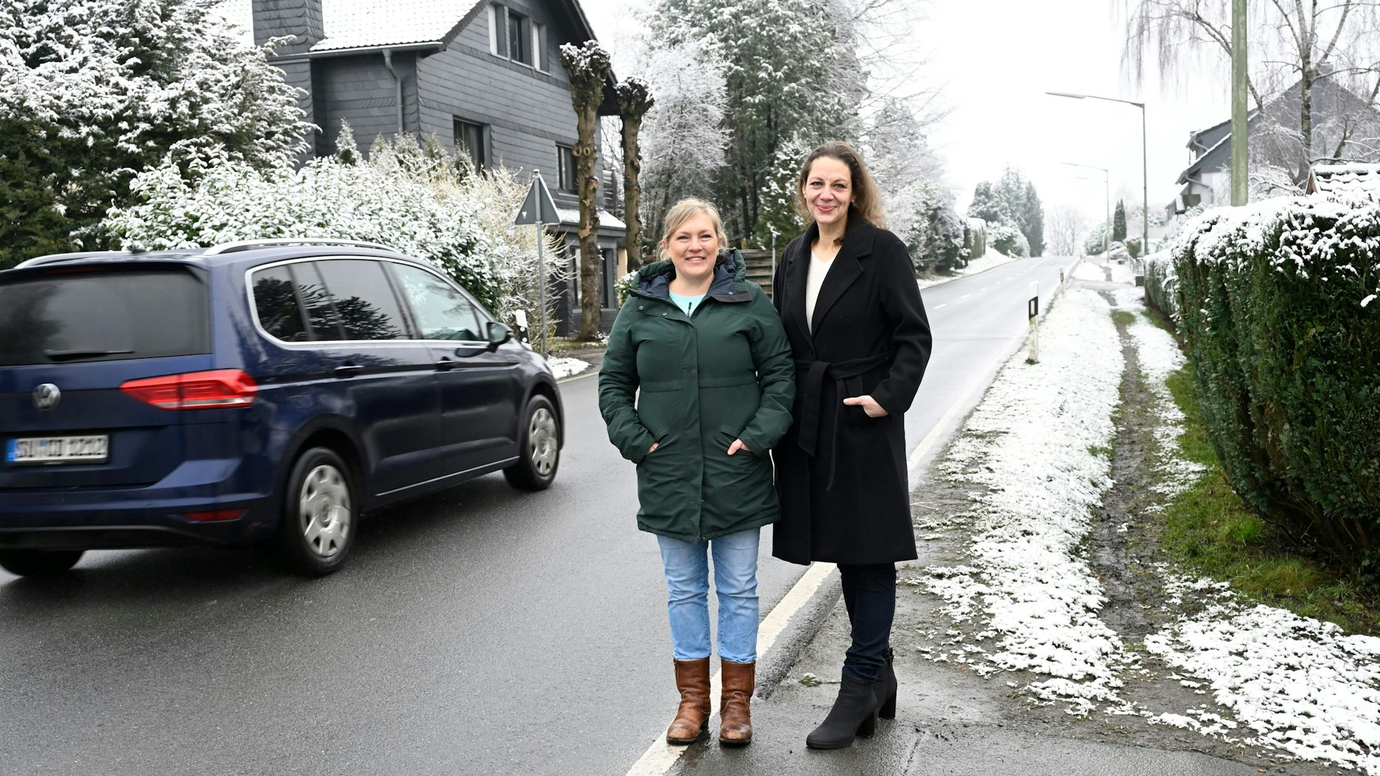 Zwei Frauen stehen neben einer Straße während ein Auto an ihnen vorbeifährt und lächeln in die Kamera.