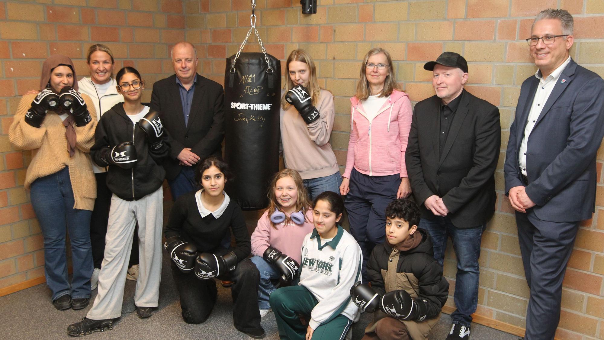 An der Gesamtschule Neunkirchen läuft seit November 2024 erfolgreich das Wahlangebot Profil Boxen. Trainierin Nikki Adler (Zweite von links) und Unterstützer Joey Kelly (Zweiter von rechts) wehten den neuen Boxsack mit Schulleiter Alfred Himpeler (Vierter von links) und Bürgermeister Guido Vierkötter (rechts) ein.