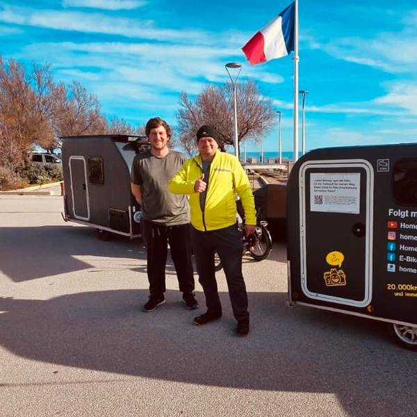 Zwei Männer stehen neben ihren Fahrradcampern auf der Promenade der südfranzösischen Hafenstadt Sète, französische und Kölner Flagge.