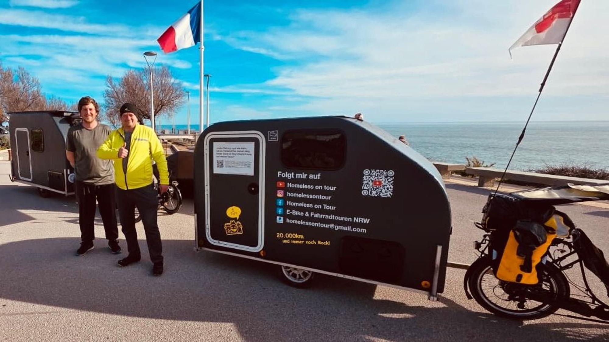 Zwei Männer stehen neben ihren Fahrradcampern auf der Promenade der südfranzösischen Hafenstadt Sète, französische und Kölner Flagge.