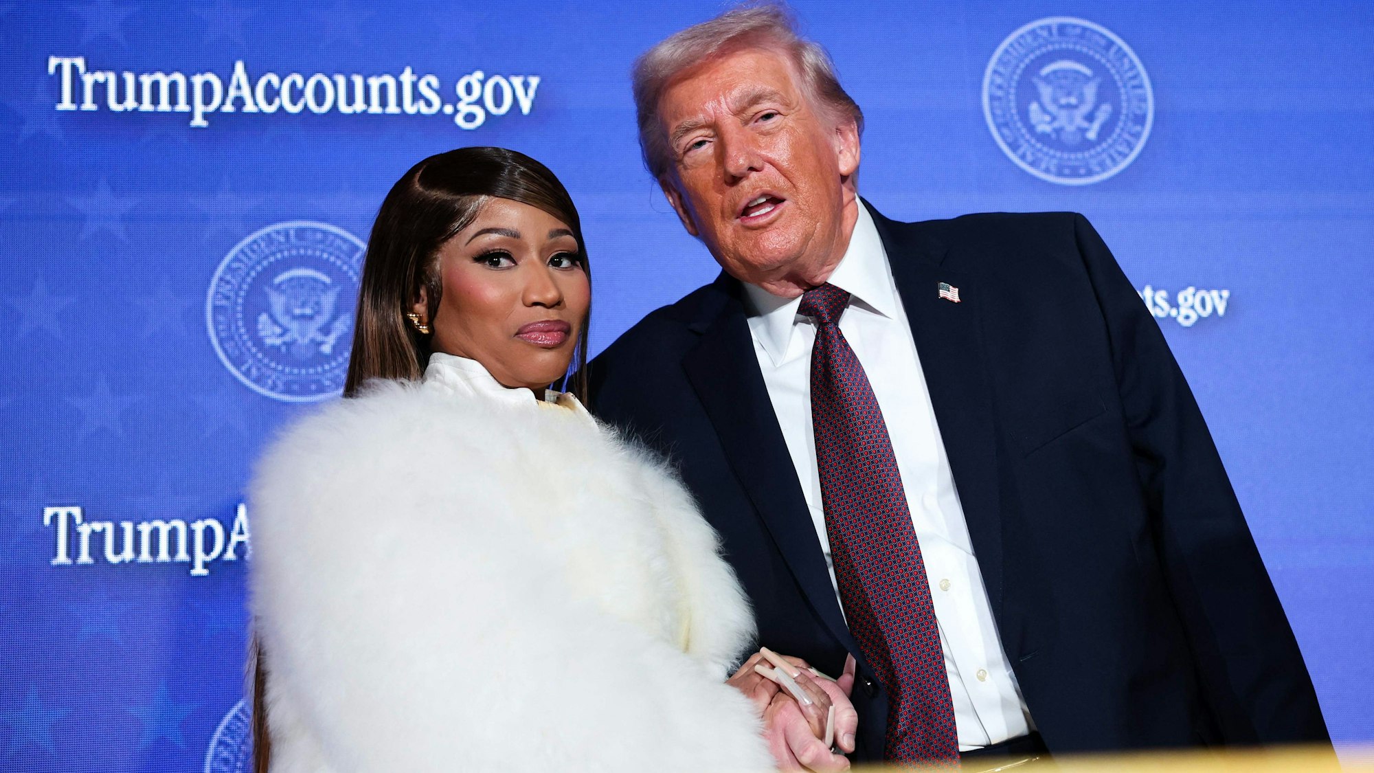 Die Musikerin Nicki Minaj (links, in weißem Kunstpelz) begleitet US-Präsident Donald Trump auf die Bühne, während er am 28. Januar 2026 im Andrew W. Mellon Auditorium in Washington, D.C., eine Rede auf dem Trump Accounts Summit des Finanzministeriums hält.