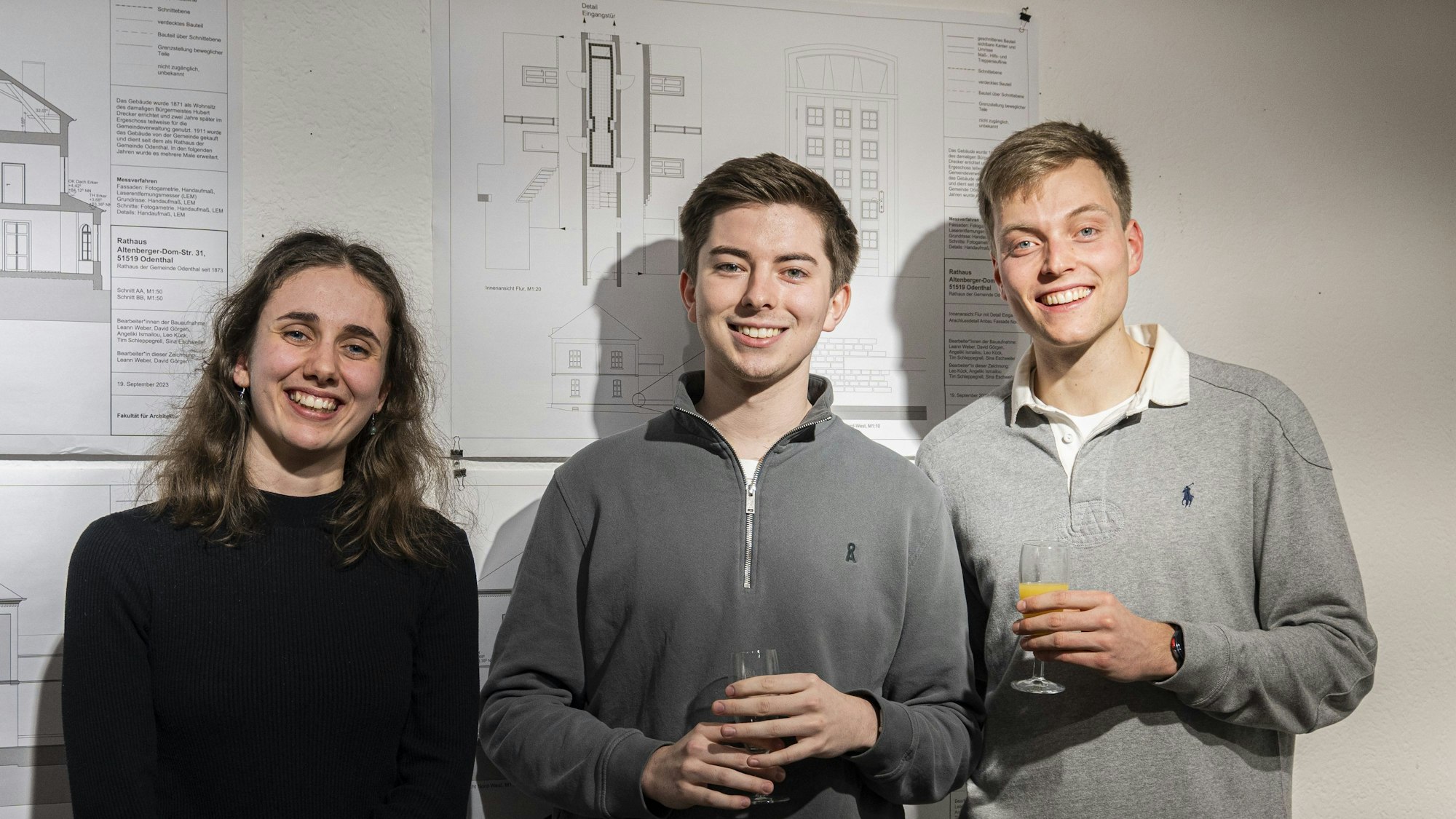 Eine junge Frau und zwei Männer stehen vor Bauplänen.