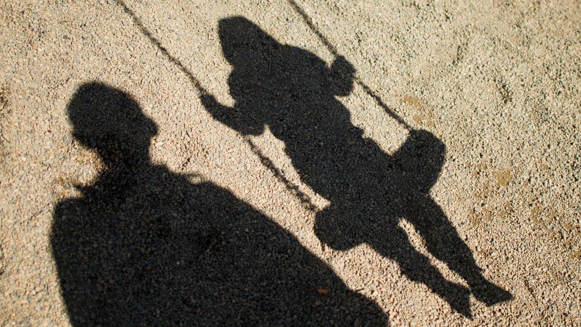 Der Schatten von einem Mann und einem schaukelnden Kind fallen auf Sand auf einem Spielplatz. (Symbolbild)