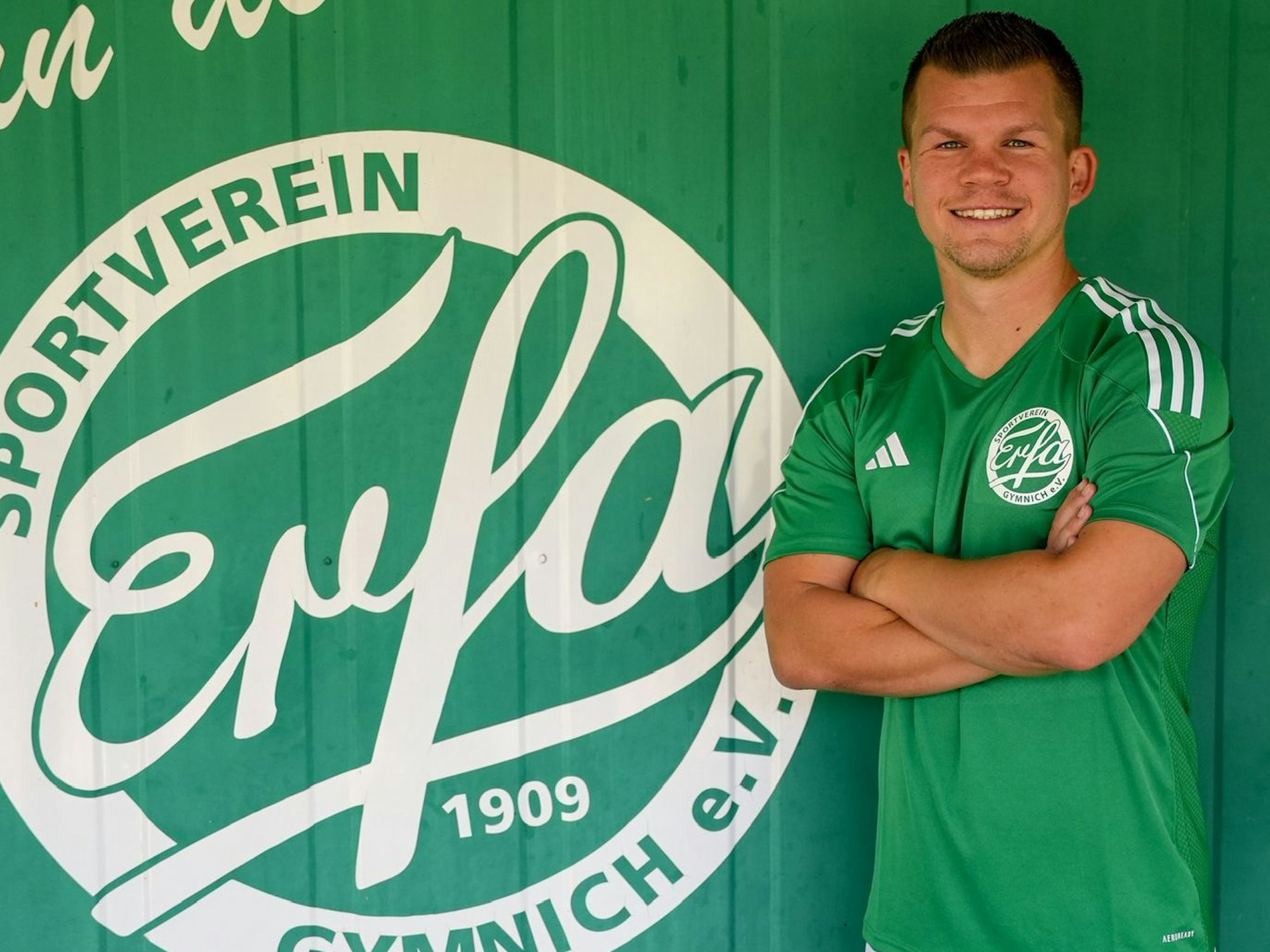 Das Bild zeigt Tobias Berg im Trikot von Erfa Gymnich, der neben dem Vereinswappen in die Kamera lächelt.