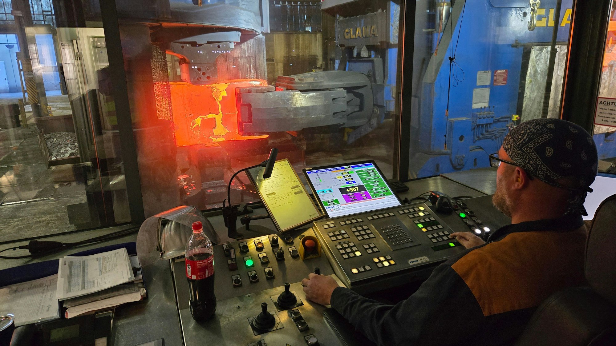 Ein Mann an einem Steuerungspult, dahinter ein glühender Block Stahl in einer Schmiedepresse.