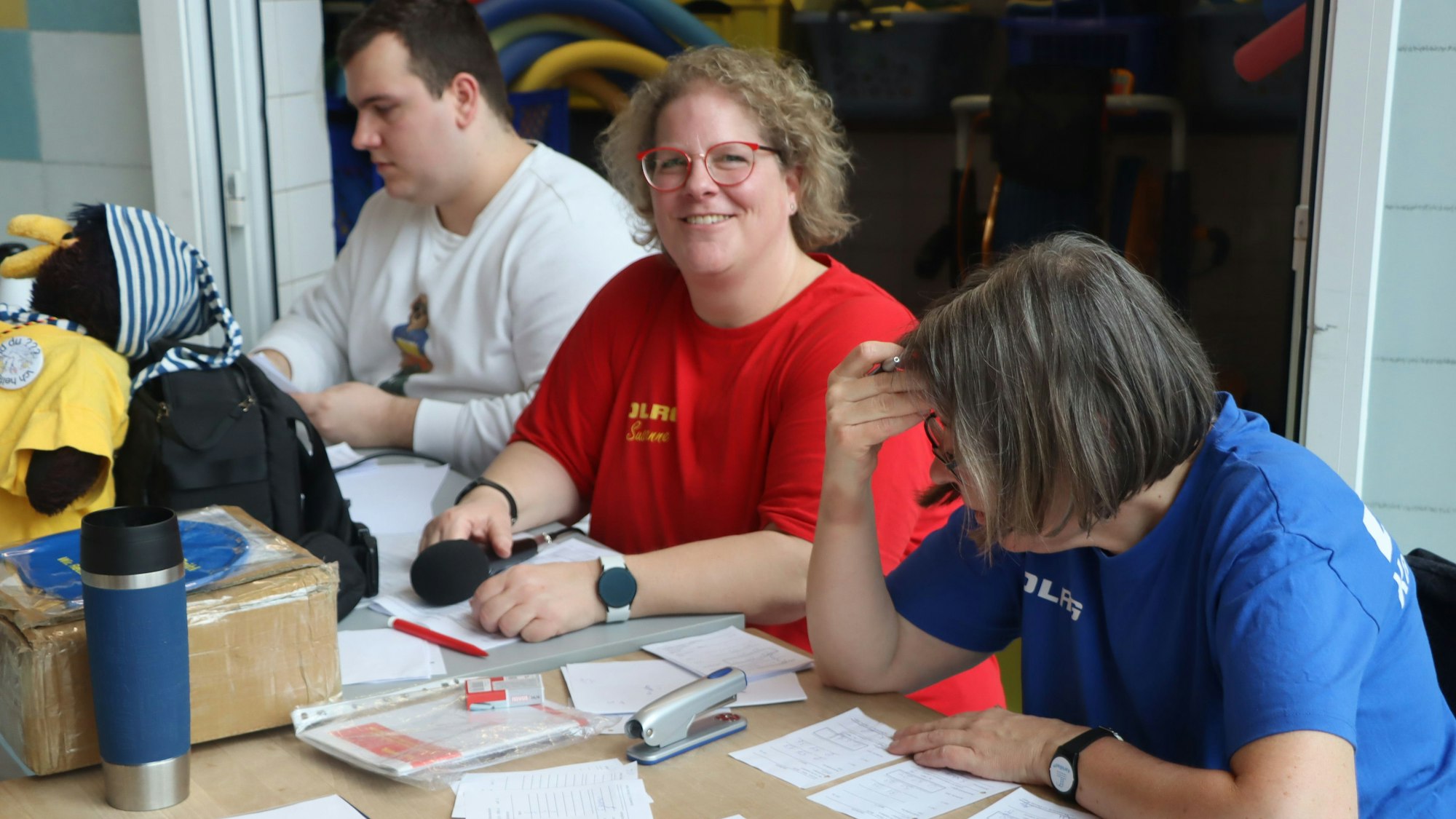 Zu sehen sind zwei Frauen und ein Mann, die an einem Tisch voller Listen sitzen.