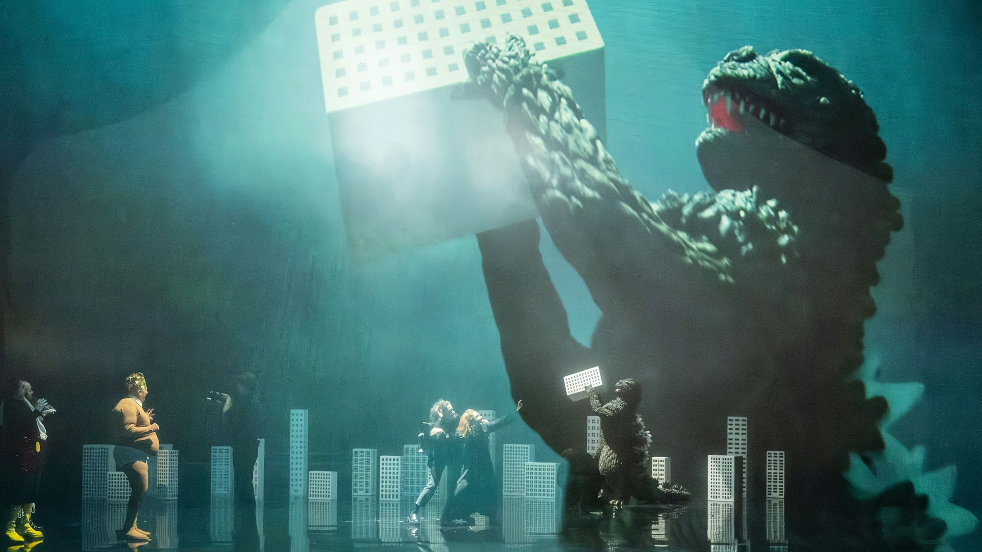 Auf einer Opernbühne hebt ein Monster ein Gebäude hoch.