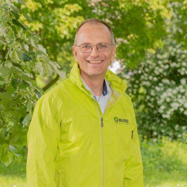 Dirk Jansen trägt eine Brille und eine hellgrüne Jacke. Er lächelt in die Kamera. Im Hintergrund Bäume und Büsche.