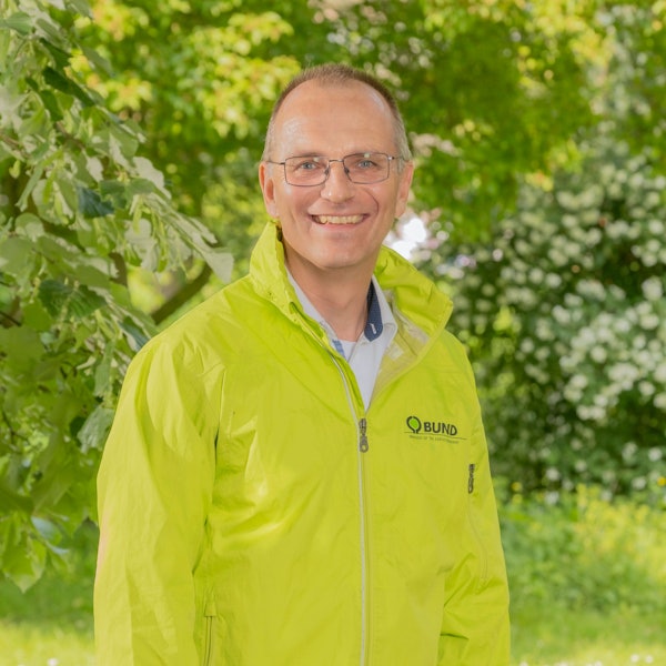Dirk Jansen trägt eine Brille und eine hellgrüne Jacke. Er lächelt in die Kamera. Im Hintergrund Bäume und Büsche.