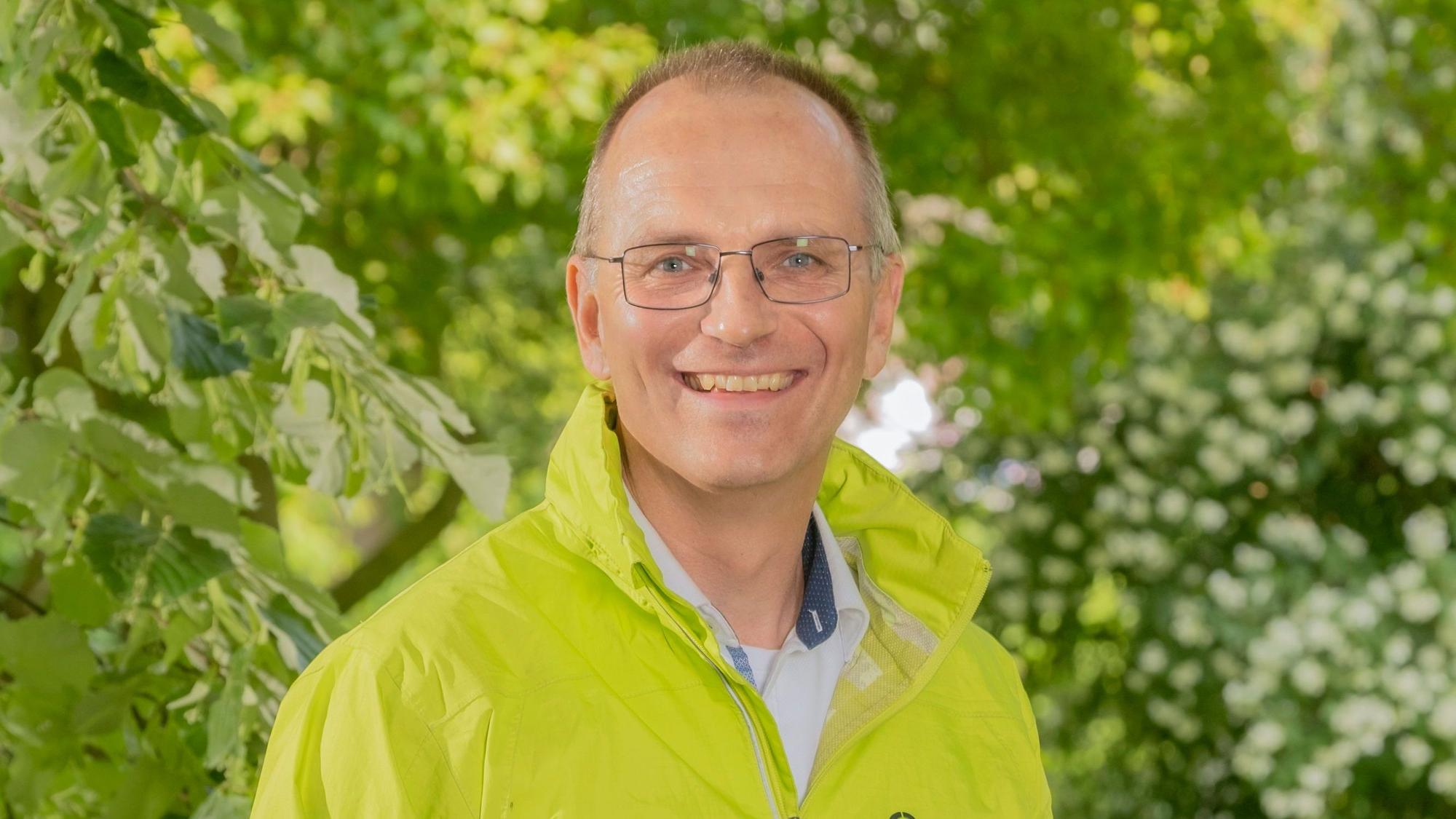 Dirk Jansen trägt eine Brille und eine hellgrüne Jacke. Er lächelt in die Kamera. Im Hintergrund Bäume und Büsche.