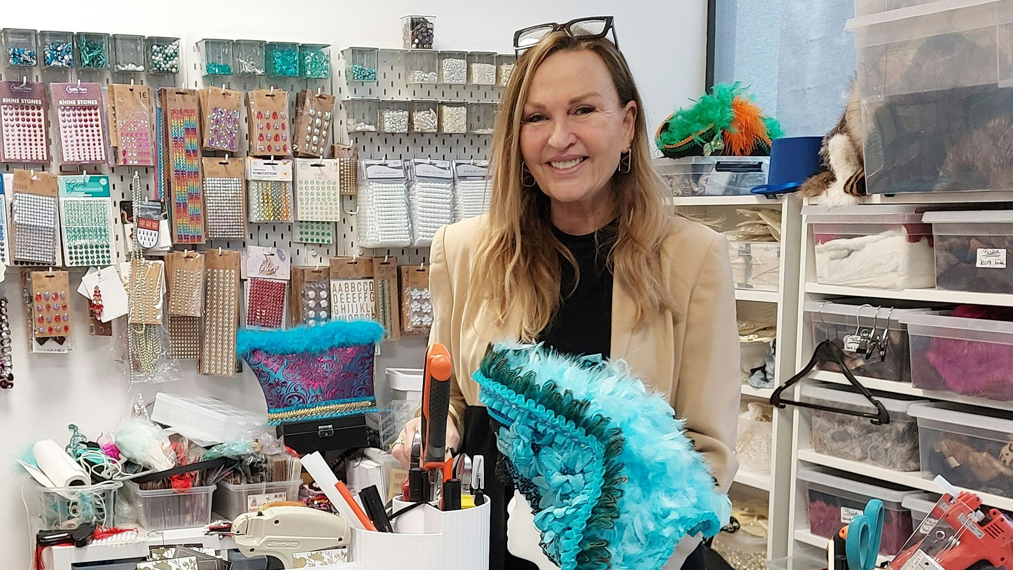 Eine Frau in einem Schneideratelier. Sie hat einen bunten Karnevalshut mit Federn in der Hand.