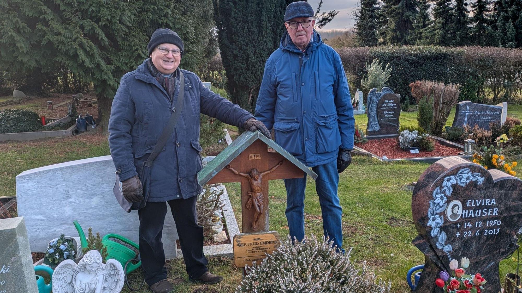 Zwei Männer stehen an einem Grab mit einem Holzkreuz.