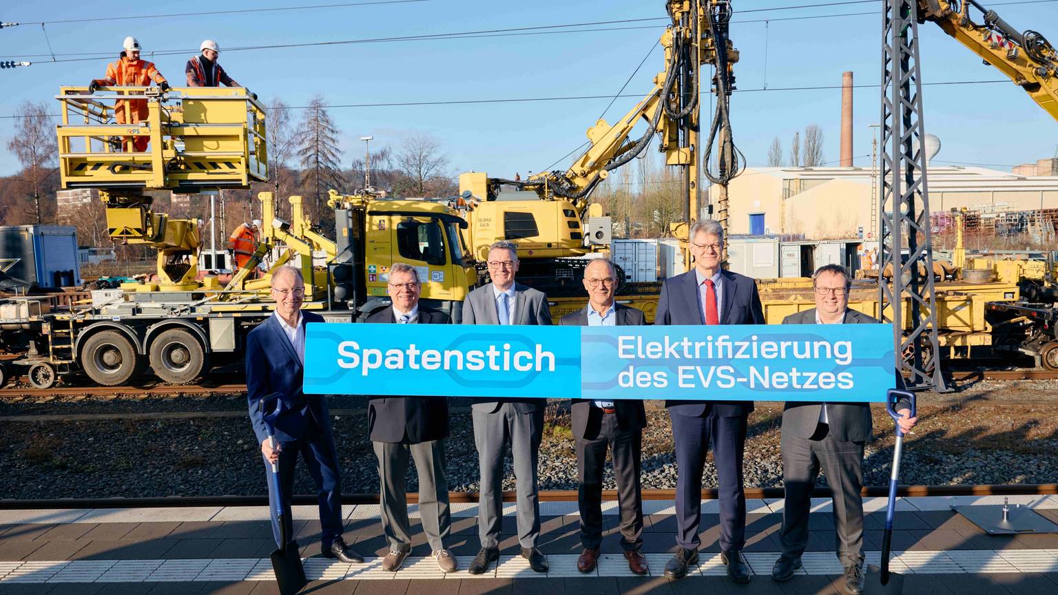 Die Herren halten ein Transparent mit der Aufschrift "Spatenstich" hoch.