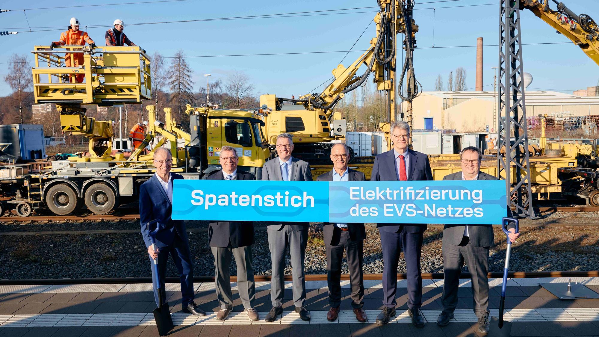 Die Herren halten ein Transparent mit der Aufschrift "Spatenstich" hoch.