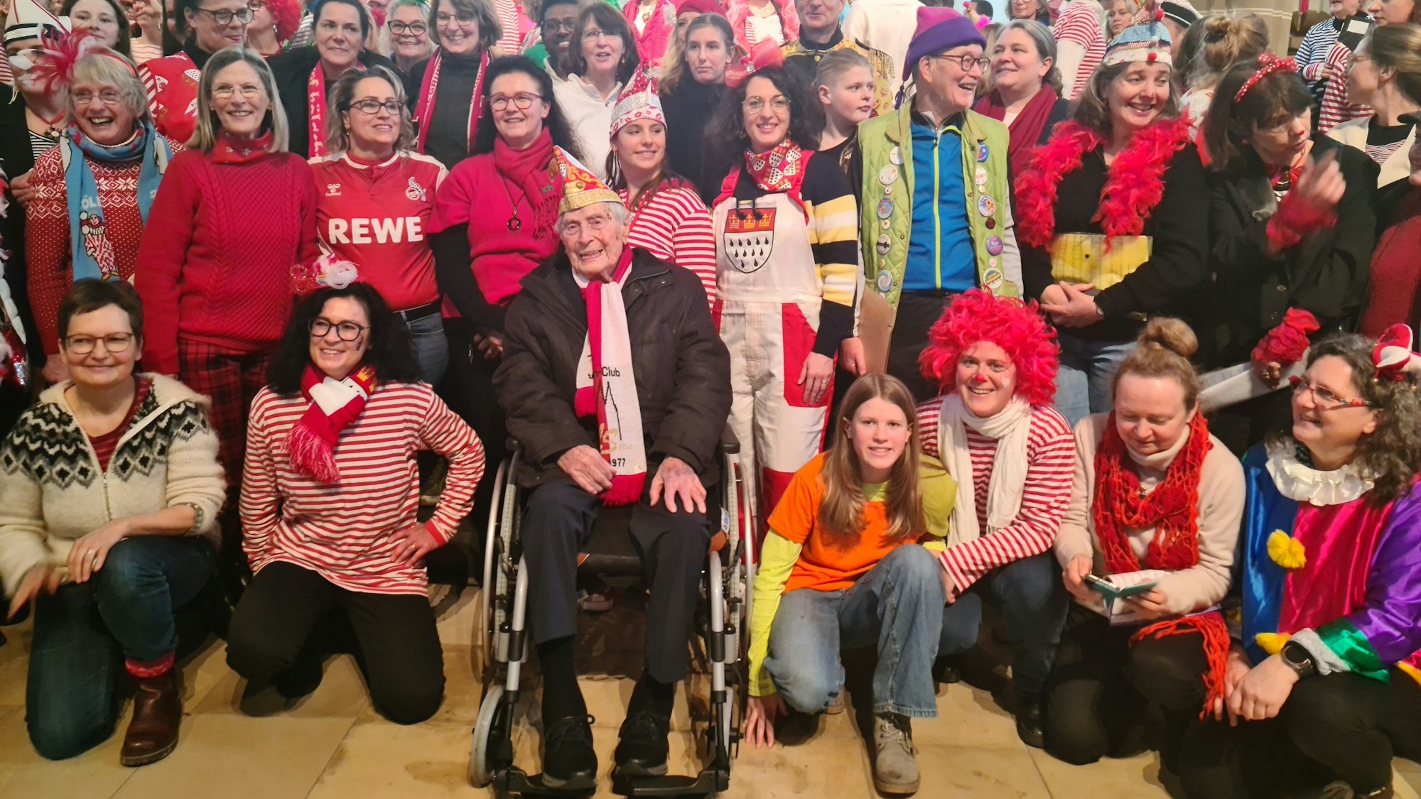 Viele kostümierte Menschen stehen in mehreren Reihen in einer Kirche, in der Mitte im Rollstuhl ein alter Herr im Anzug mit Köln-Schal.