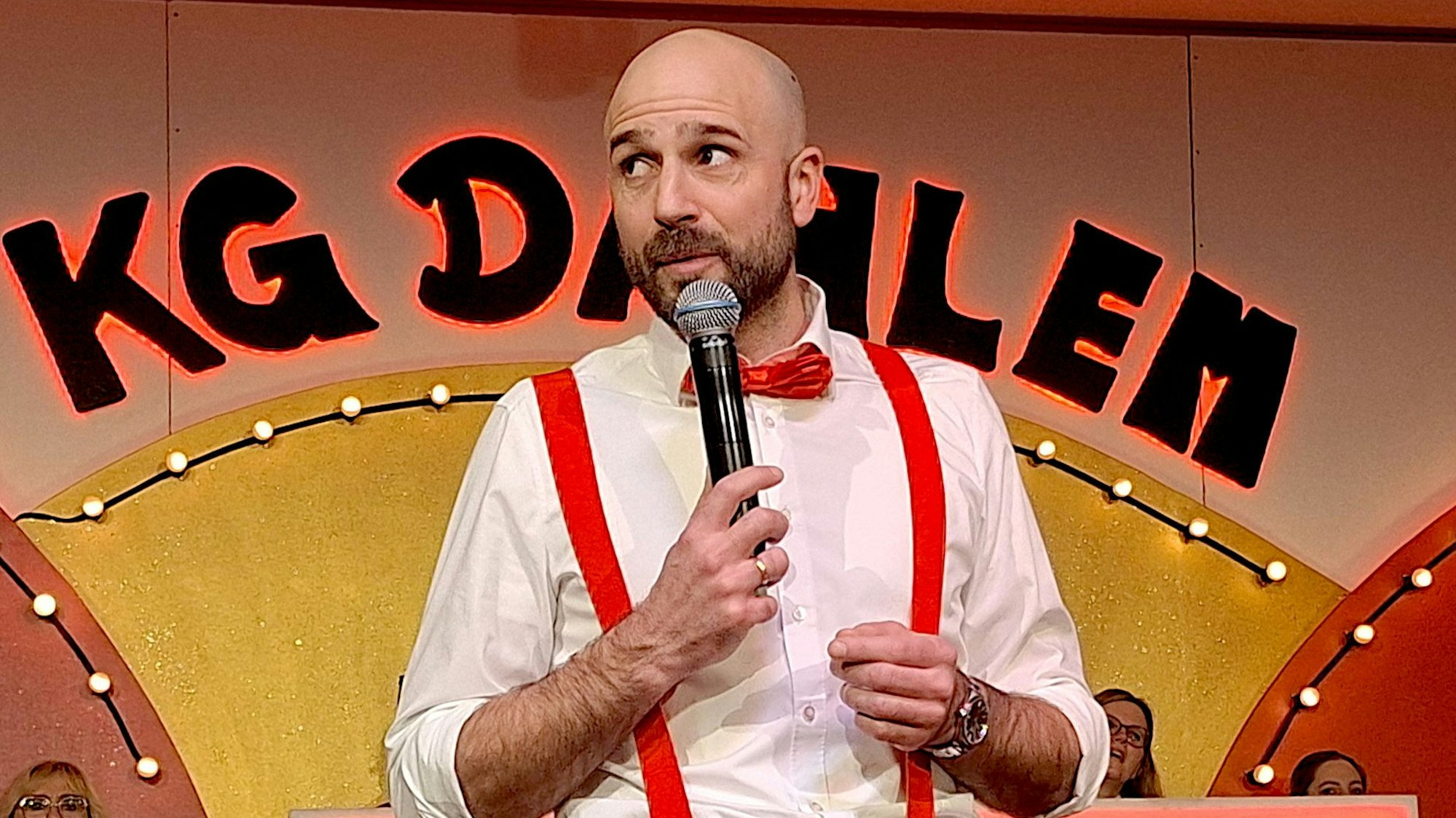 Daniel Klinkhammer, im weißen Hemd und mit roten Hosenträgern, steht mit einem Mikrofon in der Hand auf der Bühne.