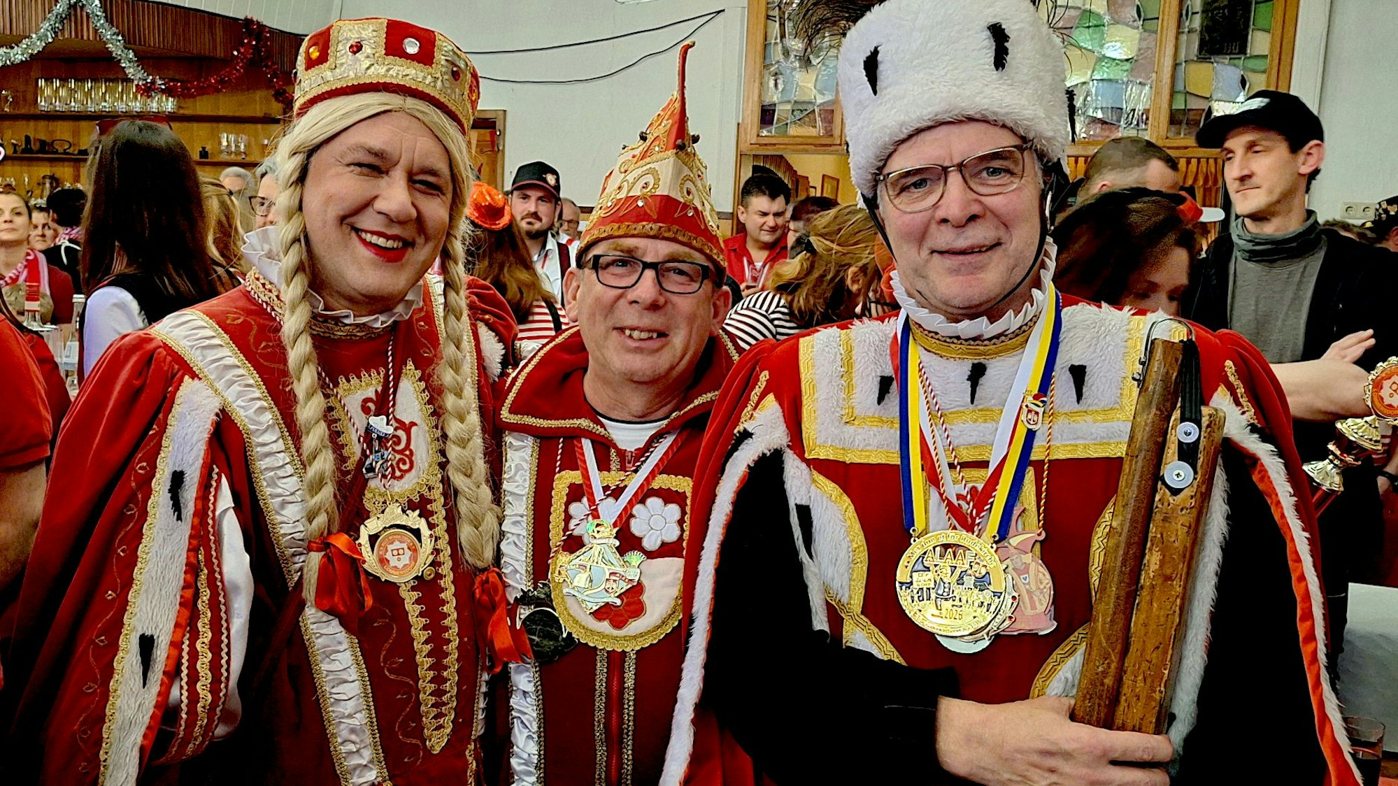 Die drei Männer des Kommersdorfer Dreigestirns stehen verkleidet und mit Orden in einem Saal voller Menschen.