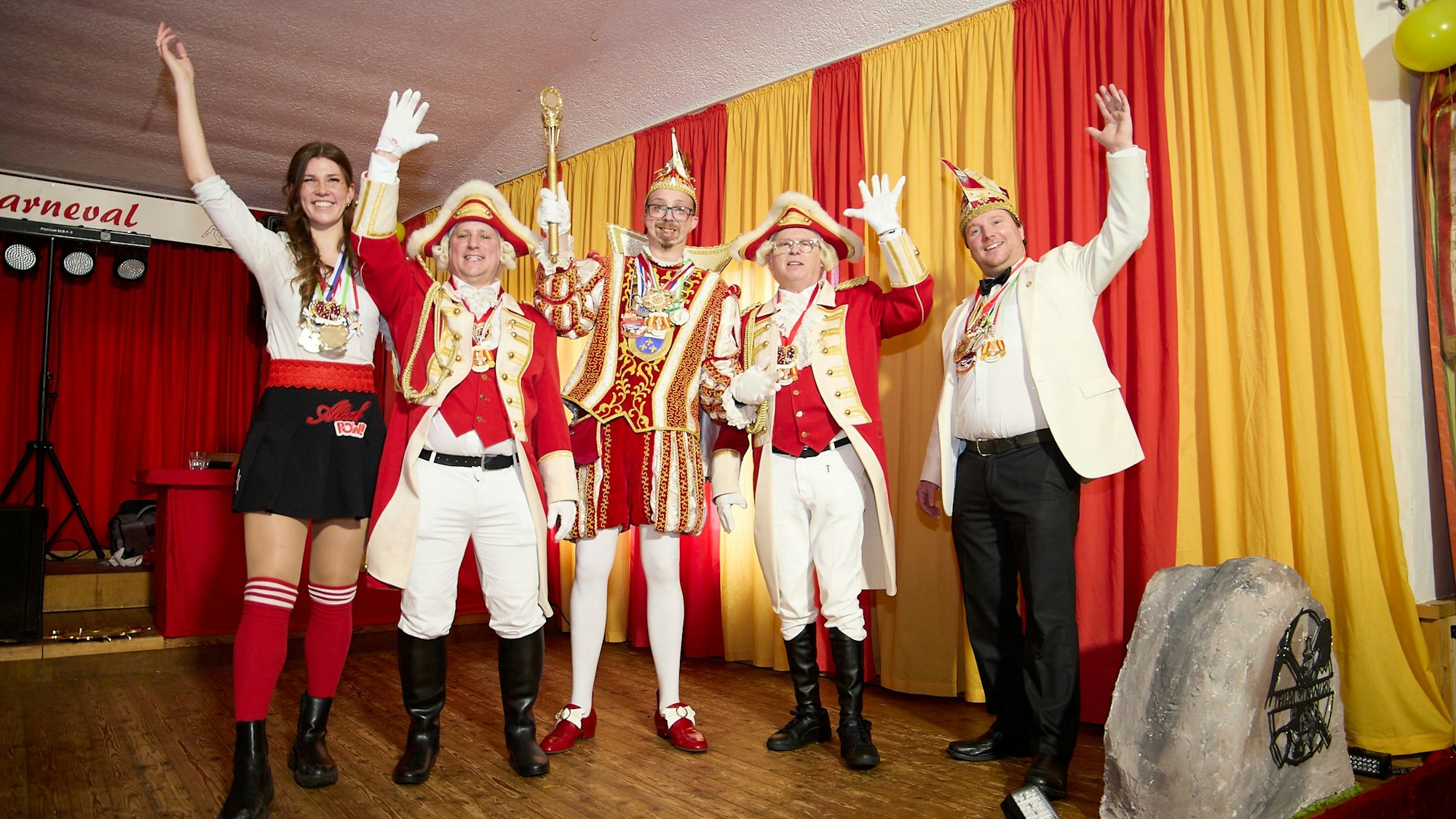 Vier Karnevalisten und eine Karnevalistin stehen auf einer Bühne und werfen jeweils eine Hand in die Luft.