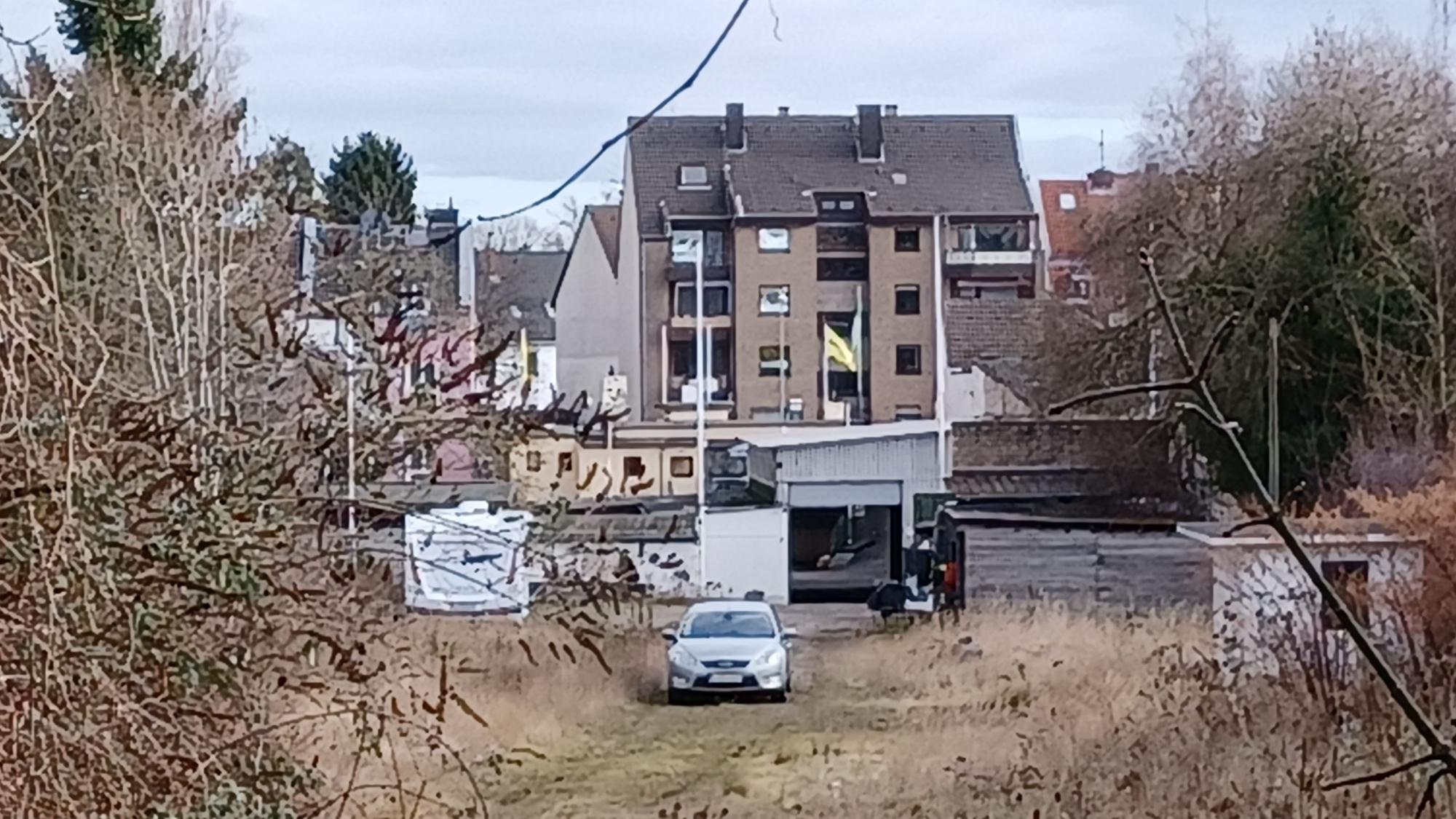 Auf einer Brachfläche steht ein Auto, im Hintergrund sind Häuser zu sehen.