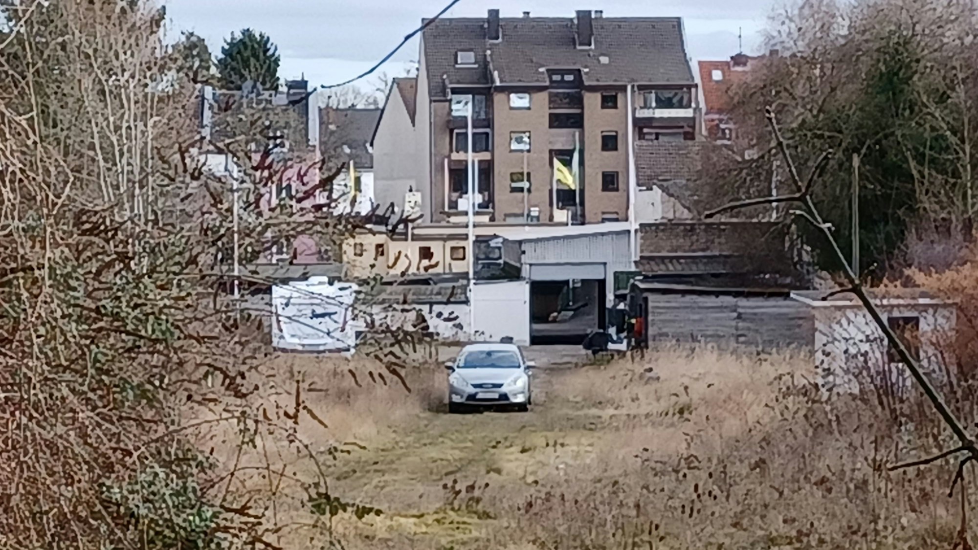 Auf einer Brachfläche mit viel Gestrüpp parkt ein einsames Auto. Im Hintergrund sind Häuser zu sehen.