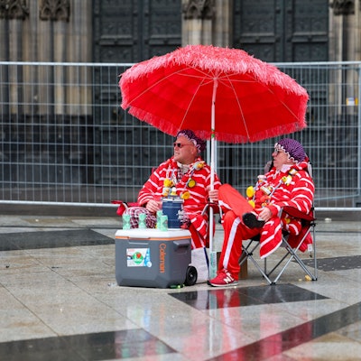 Zwei Karnevalisten sitzen bei regnerischem Wetter unter einem Sonnenschirm auf der Domplatte.