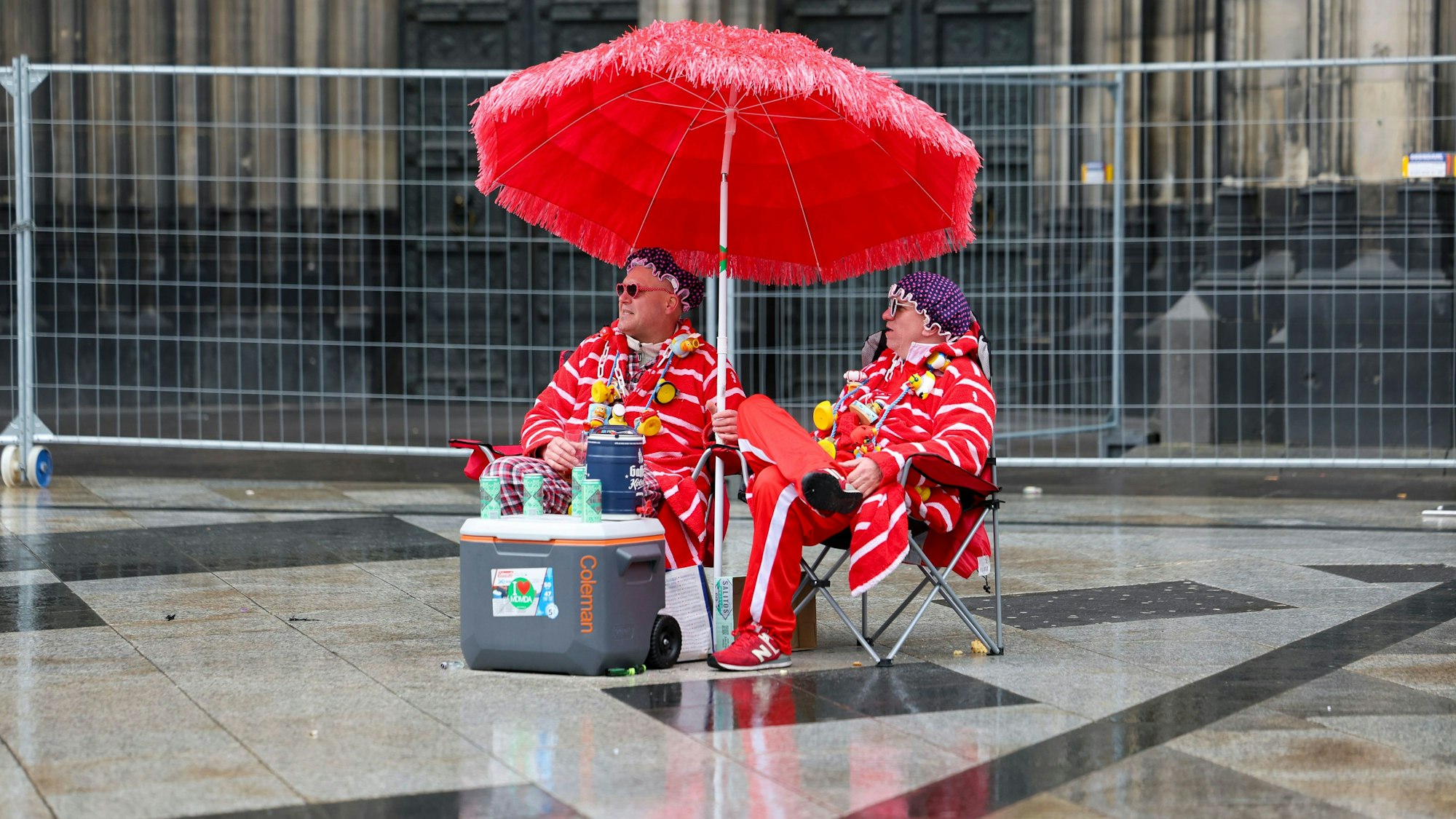 Zwei Karnevalisten sitzen bei regnerischem Wetter unter einem Sonnenschirm auf der Domplatte.