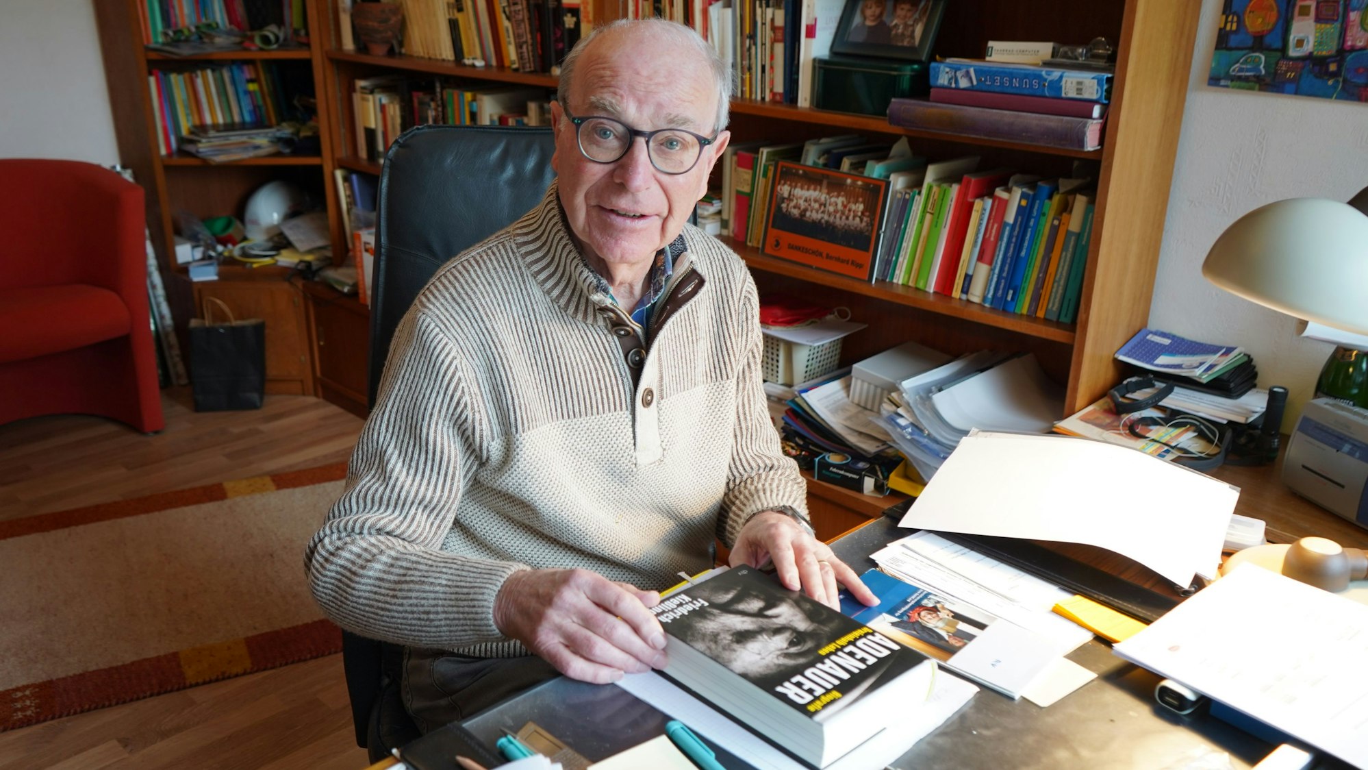 Das Foto zeigt einen älteren Herren an einem Schreibtisch, im Hintergrund stehen Bücher im Regal.