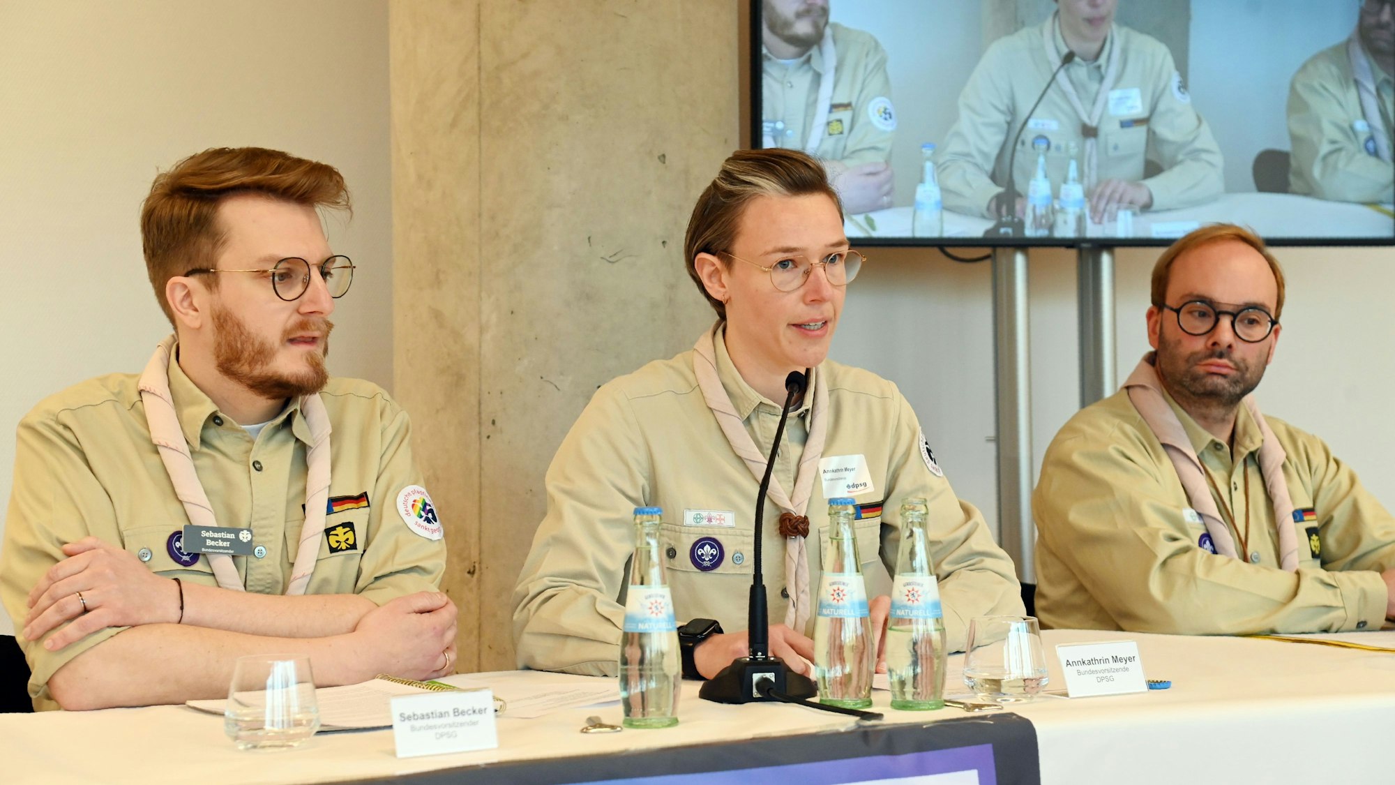 Drei Personen sitzen nebeneinander an einem Tisch und geben eine Pressekonferenz.