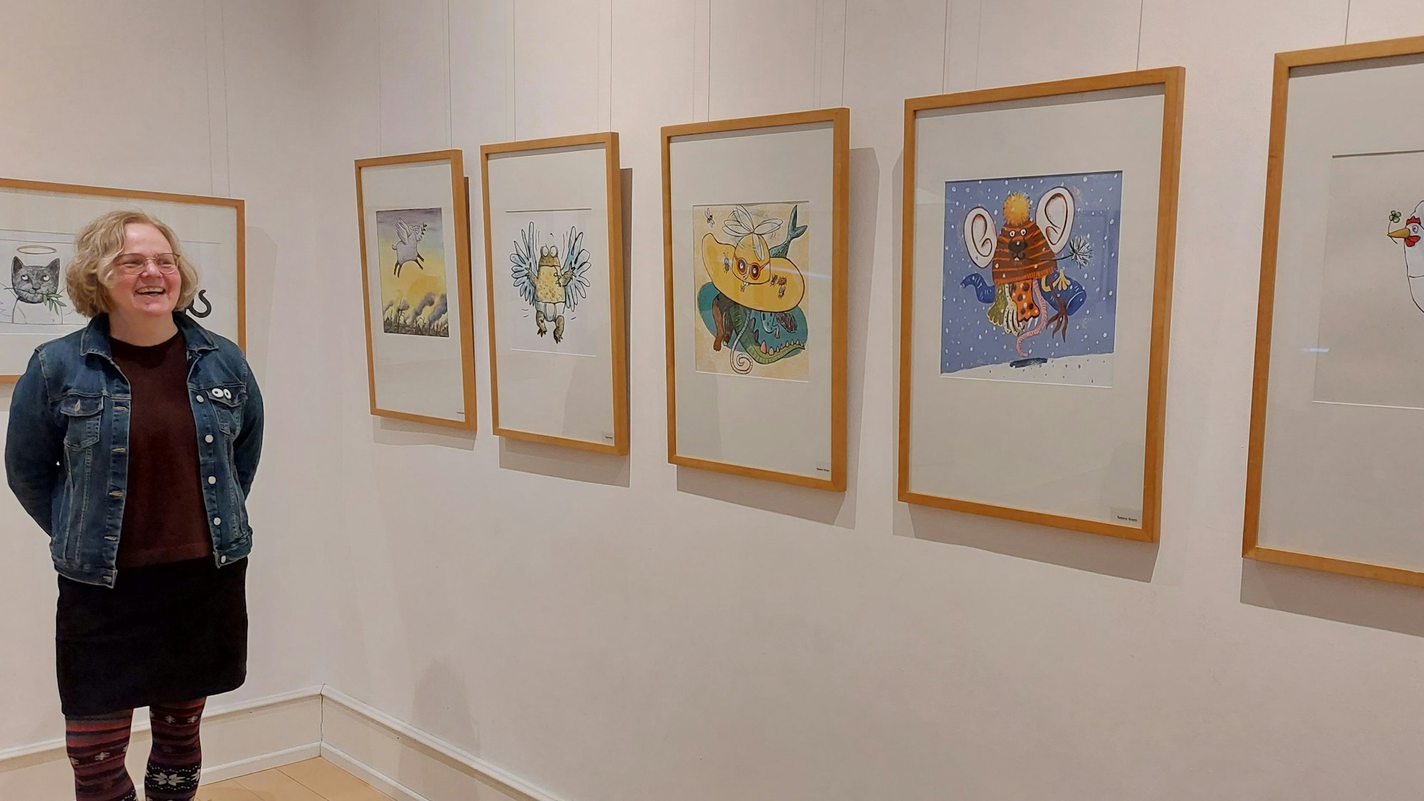 Eine Frau mit blonden Locken steht neben einer Museumswand mit gerahmten Illustrationen.