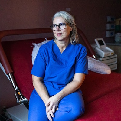 Eine Frau im blauen Kasack schaut aus dem Fenster. Sie sitzt auf einem Bett im Kreissaal.