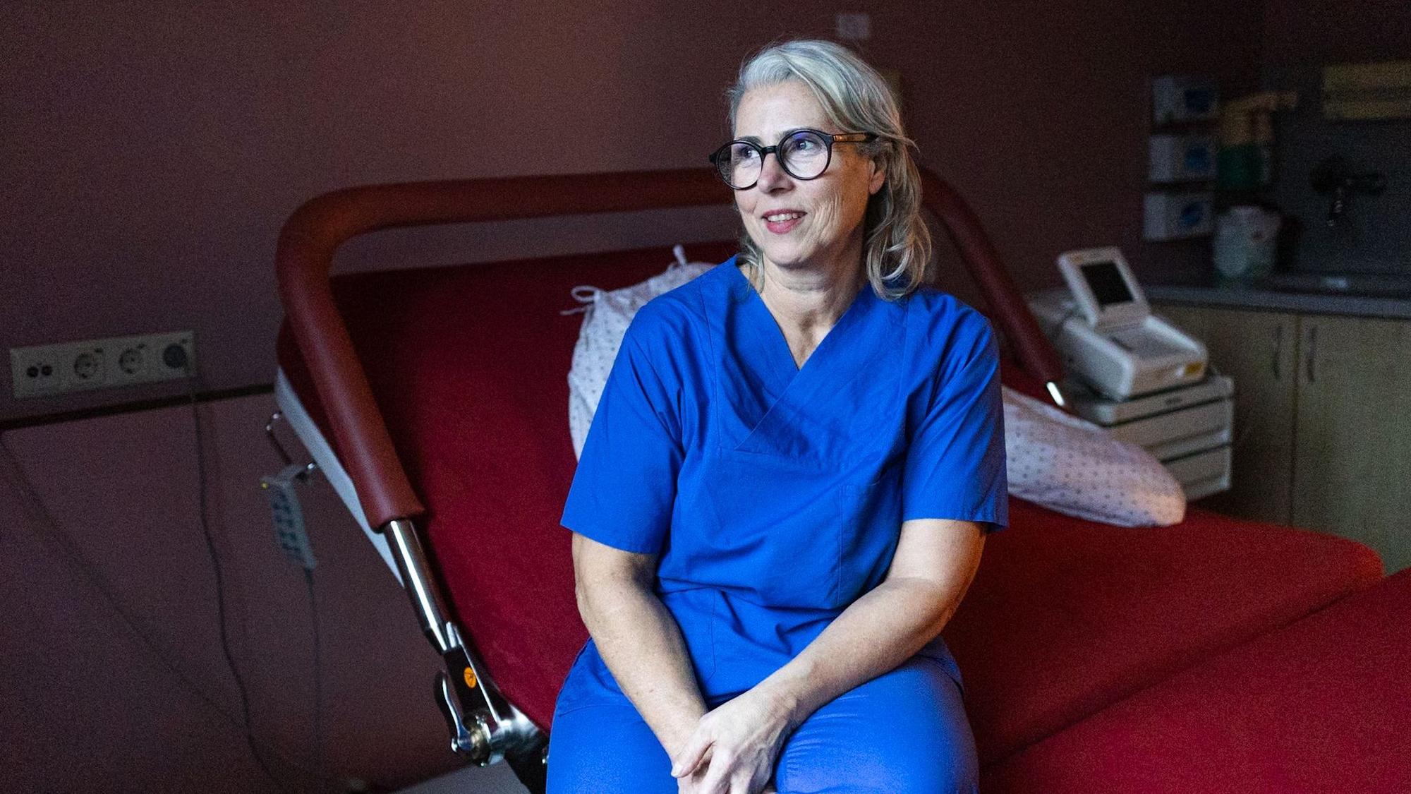 Eine Frau im blauen Kasack schaut aus dem Fenster. Sie sitzt auf einem Bett im Kreissaal.