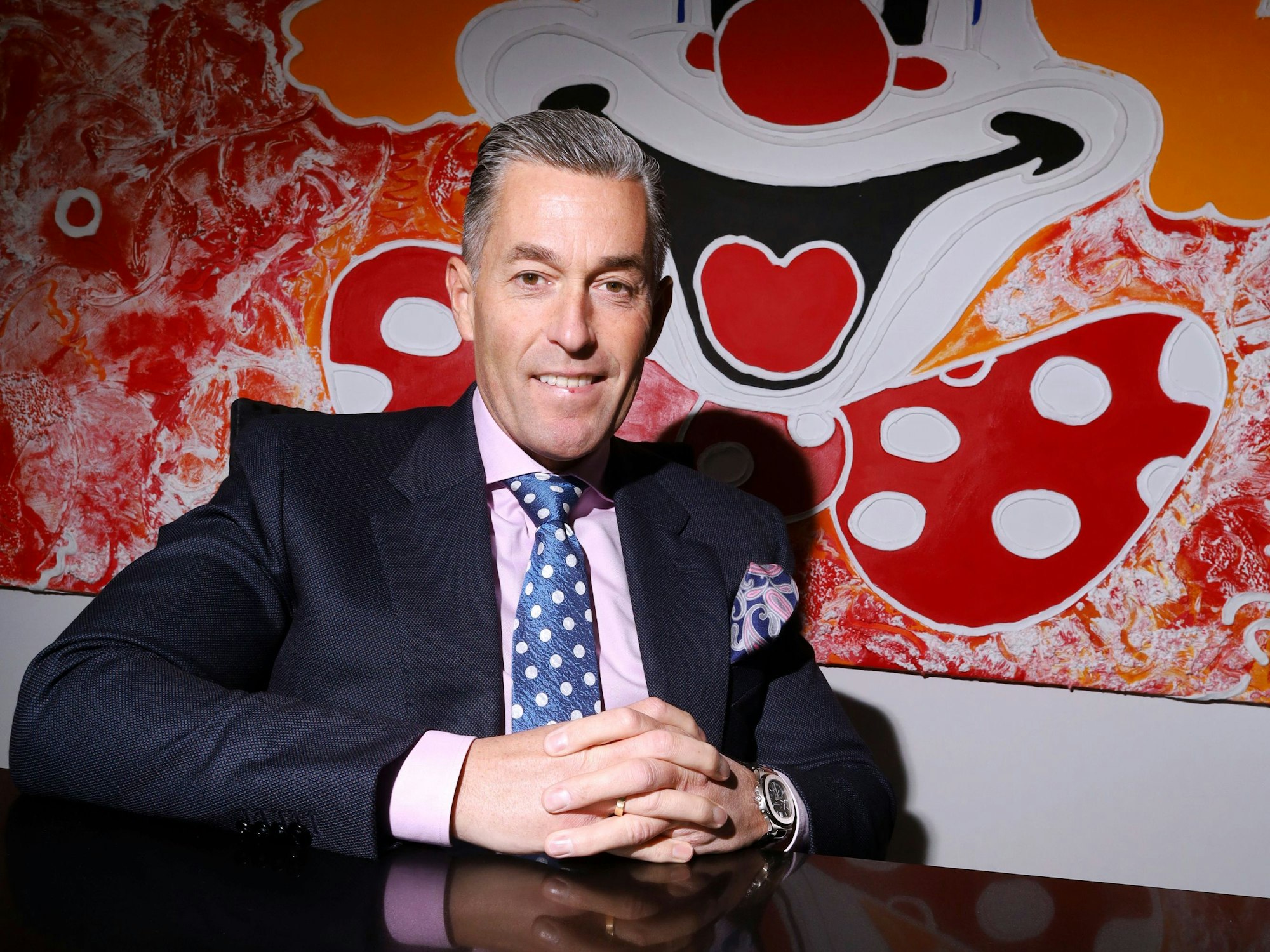 Deiters-Chef Herbert Geiss sitzt an einem schwarzen Tisch vor einem Clownsgemälde.