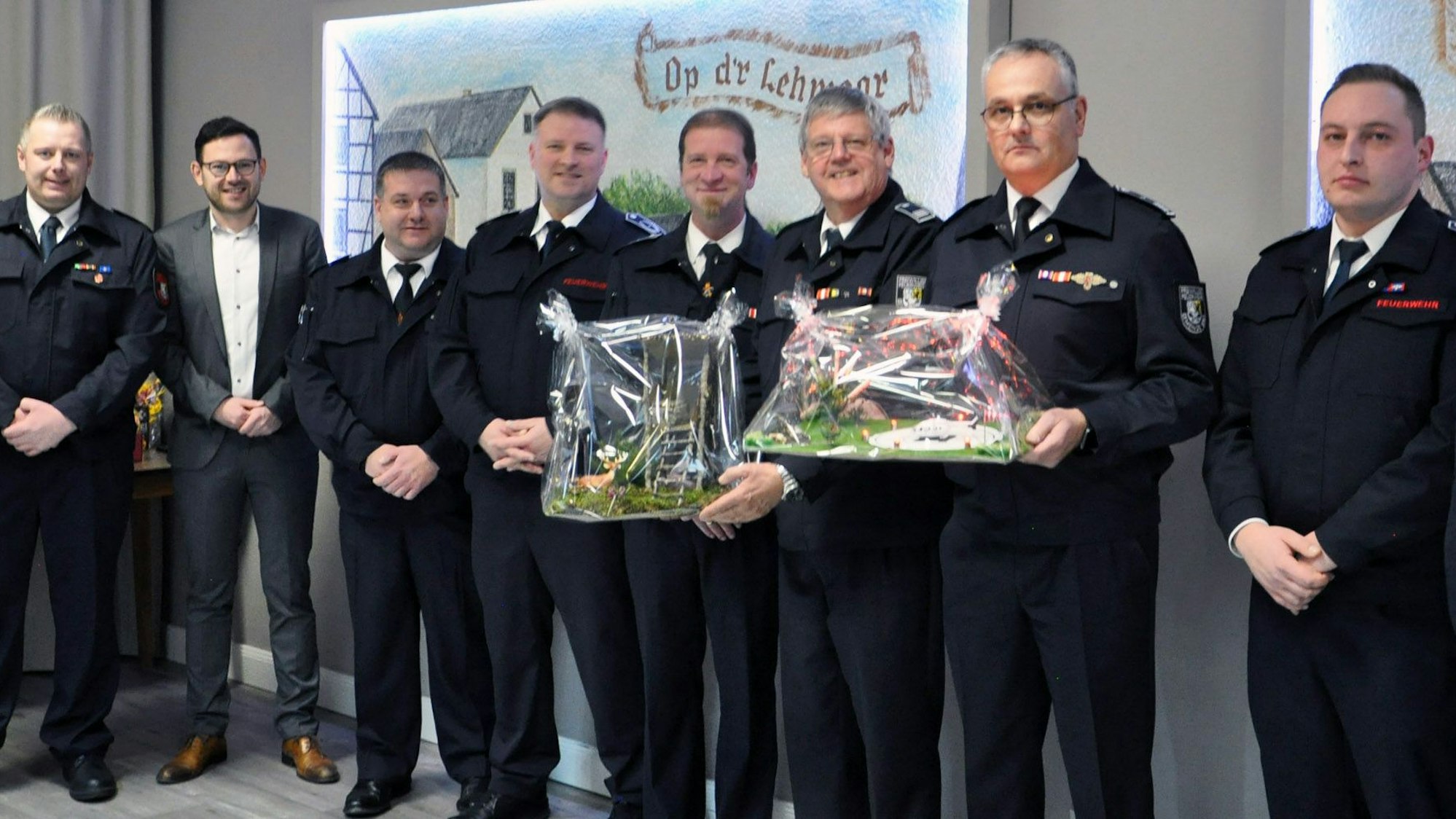Mehrere Feuerwehrmänner in Uniform udn ein Zivilist stehen nebeneinander. Zwei Männer in der Mitte halten Präsente hoch.