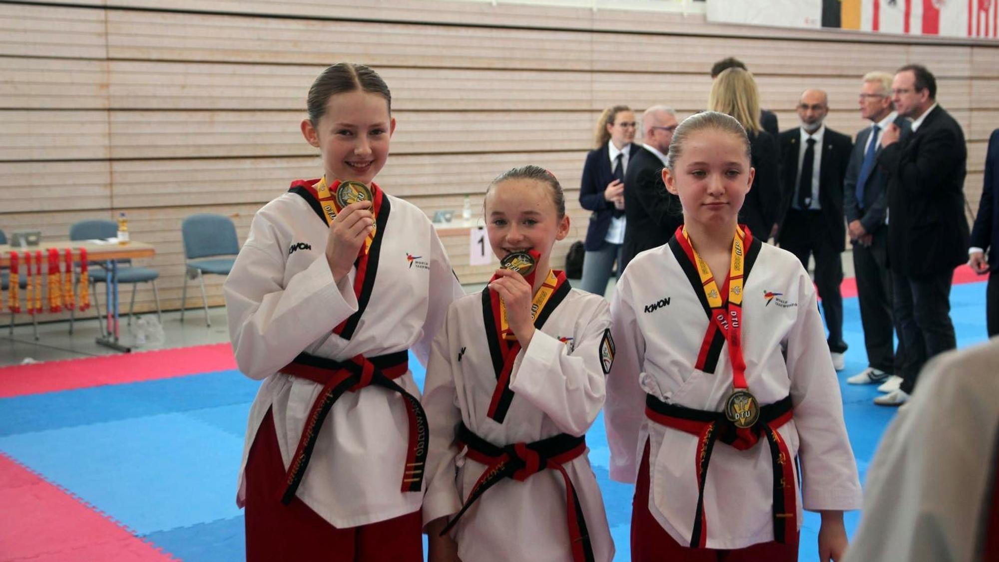 Alexandra Simin (l.) vom Taekwondo-Club Schleiden steht neben zwei anderen Sportlerinnen und hält eine Medaille in die Kamera.